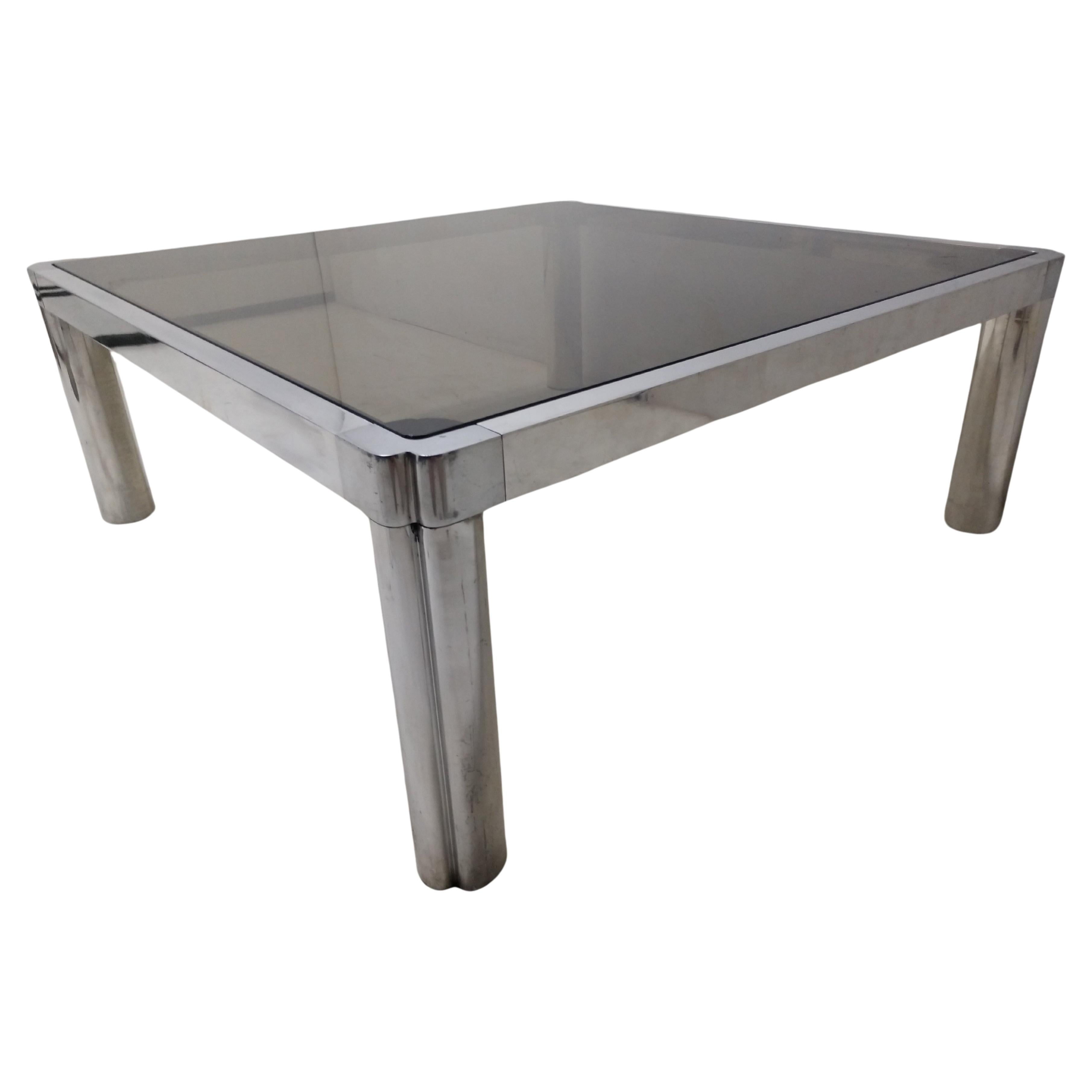 Fabulosa mesa de cóctel de aluminio fundido con tablero de cristal ahumado. Patas de tubo con barras planas que unen los lados. Pieza bien construida, ya que los herrajes son ajustados y precisos. Algunos arañazos mínimos tanto en el marco como en