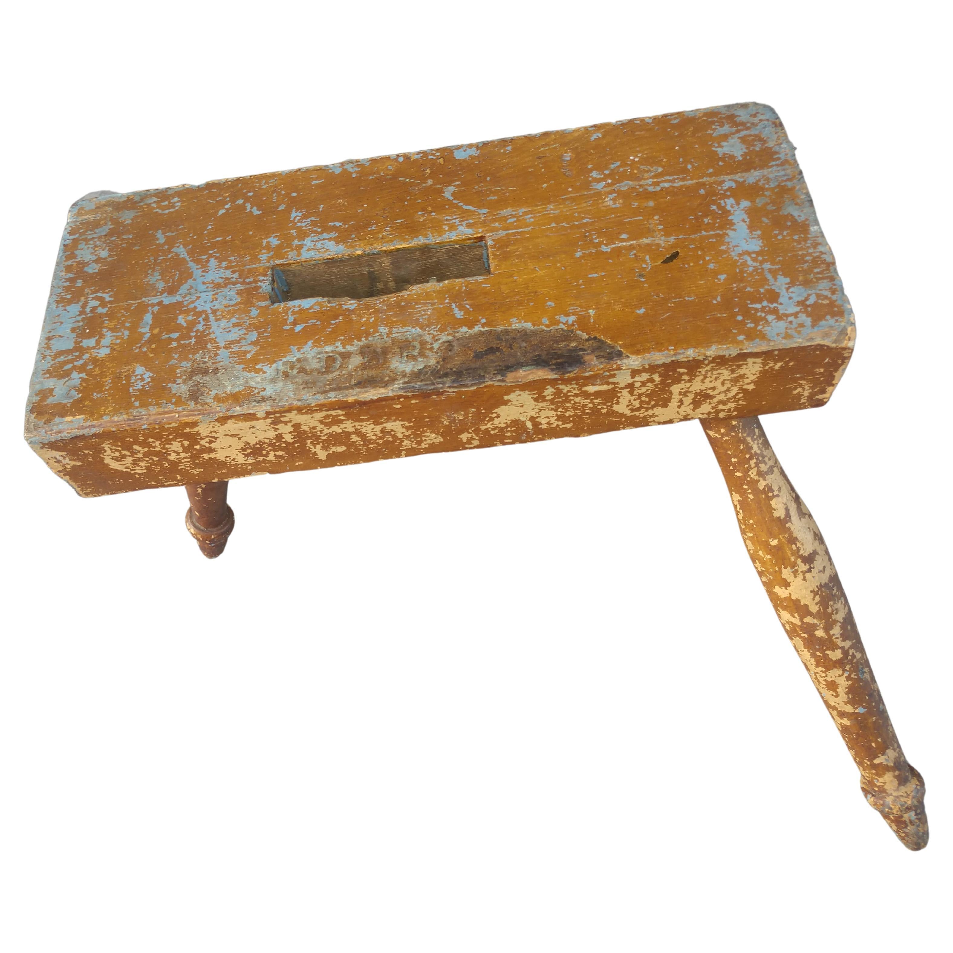 Sculptural 3 legged milking stool with multiple layers of paint. Rectangular top with a hand hole cut thru the center and supported by 3 turned legs. Their is a marking with letters on top but cannot decipher. In excellent vintage condition, very