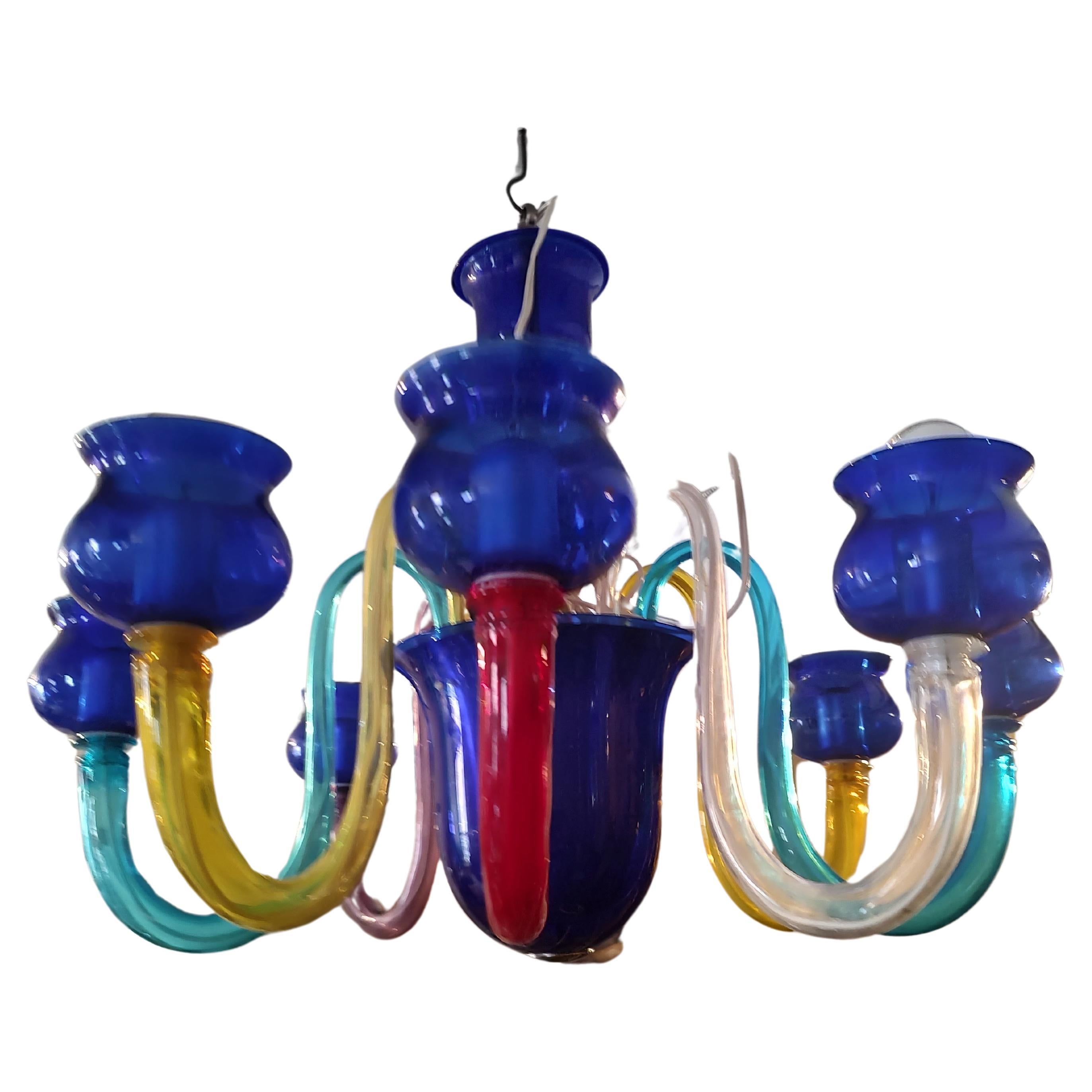 Fabuleux et élégant lustre vénitien à 8 bras de Murano. Huit bras colorés élégamment courbés, 2 rouges 2 jaunes 2 bleus 1 améthyste et 1 transparent composent le lustre avec 8 coupelles bleues qui entourent les ampoules. Des pièces centrales bleues