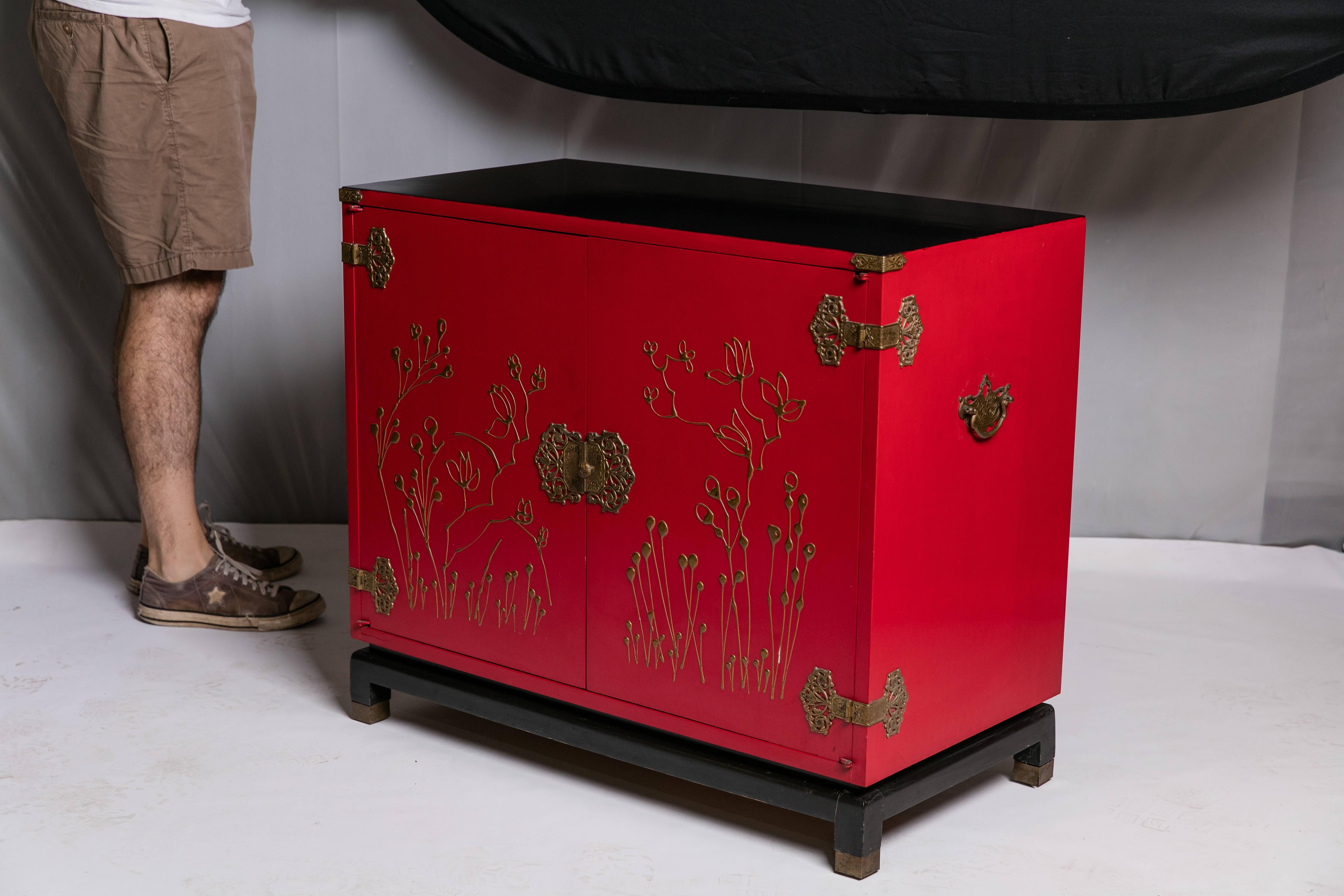 A stunning red Mid-Century wood chinoiserie bar cabinet by Bass. Styled to resemble a Chinese tea chest, the bar's red cabinet rests on raised black lacquered legs. The black top is protected by a piece of glass. The face of each door is embellished
