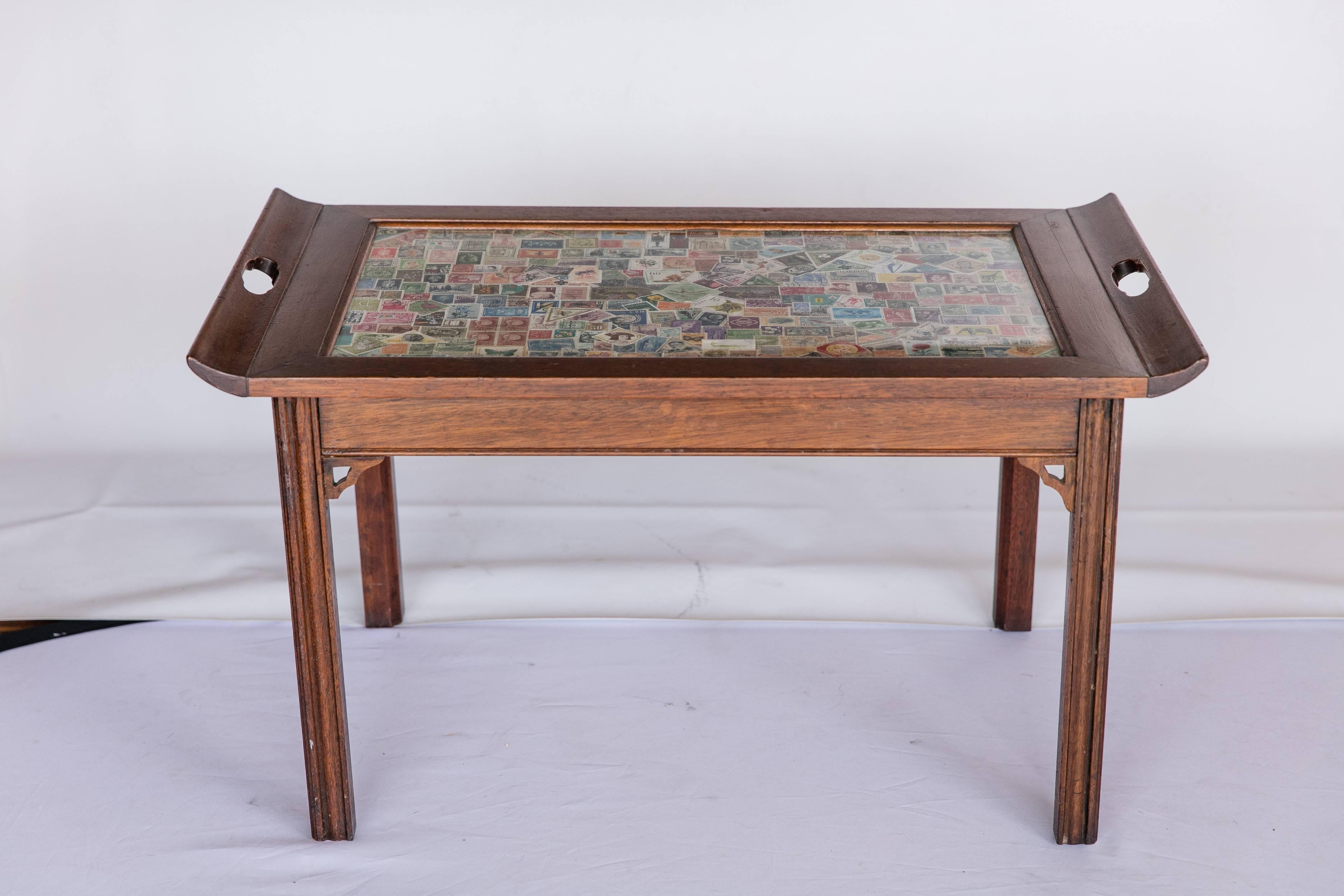 An early 20th century stained wood coffee table with top covered in a collection of international postage stamps. Stamps are protected by a glass top. Top features turned-up ends with handles for lifting and carrying table. The table is from the