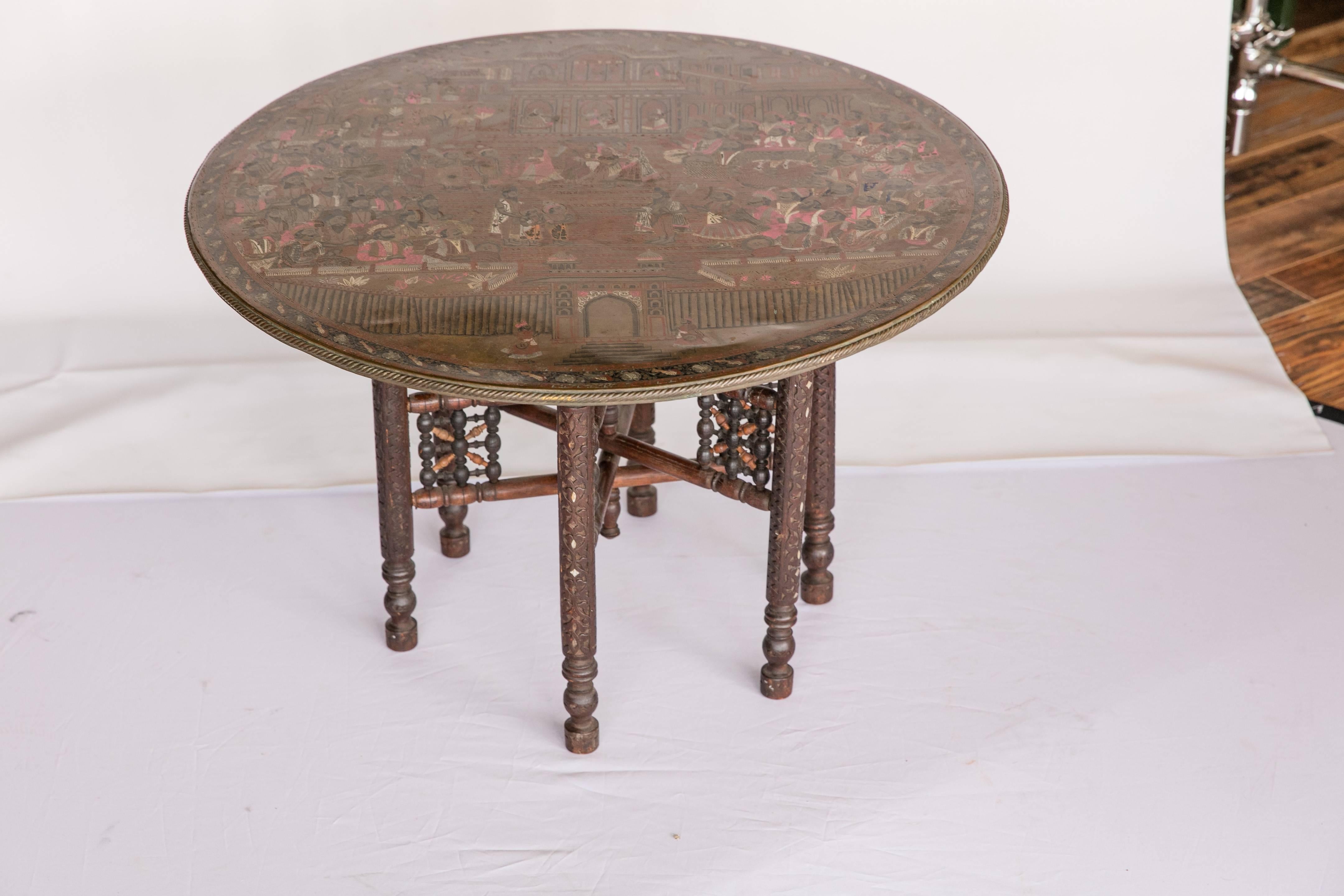 An early 20th century Indian tea table with wood base and removable brass tray top. The base features turned wood legs inlaid with mother-of-pearl. The brass tray top features an etched and painted scene of a maharajah and his court. Top can be