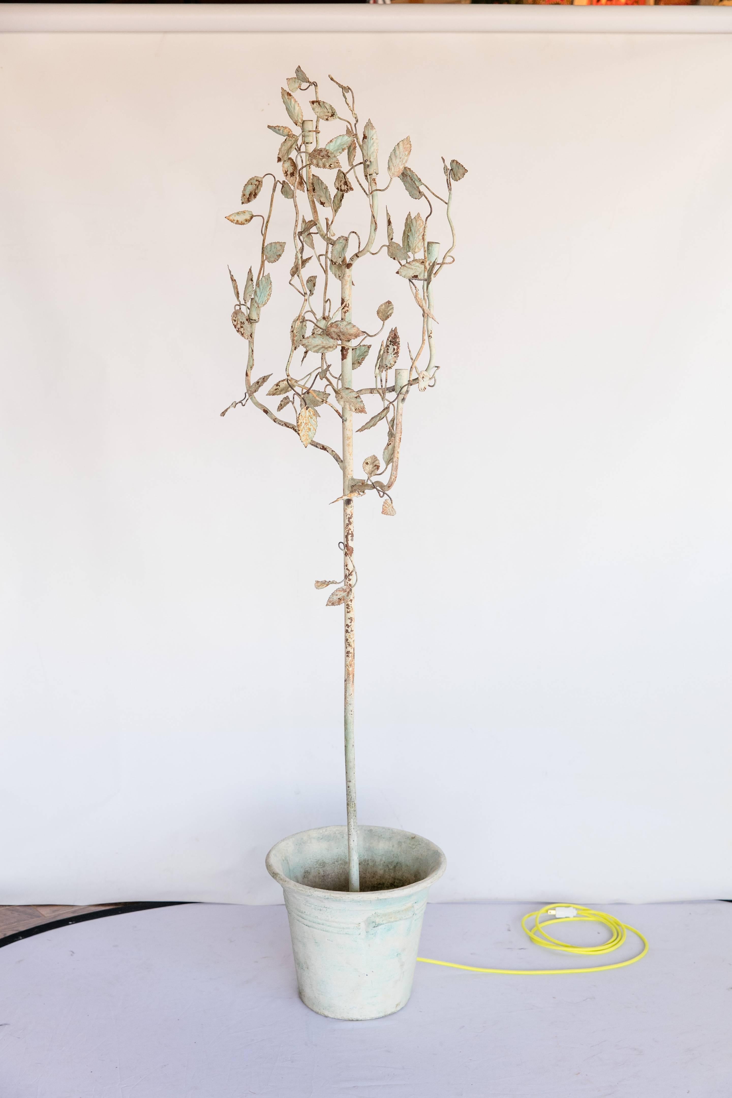A 1950s rustic metal tree floor lamp. Cream-and-light green coloration with distressing. Rust shows through distressing but adds to the overall rustic look of the piece. The tree is planted in a ceramic pot. The tree's branches conceal five lights.