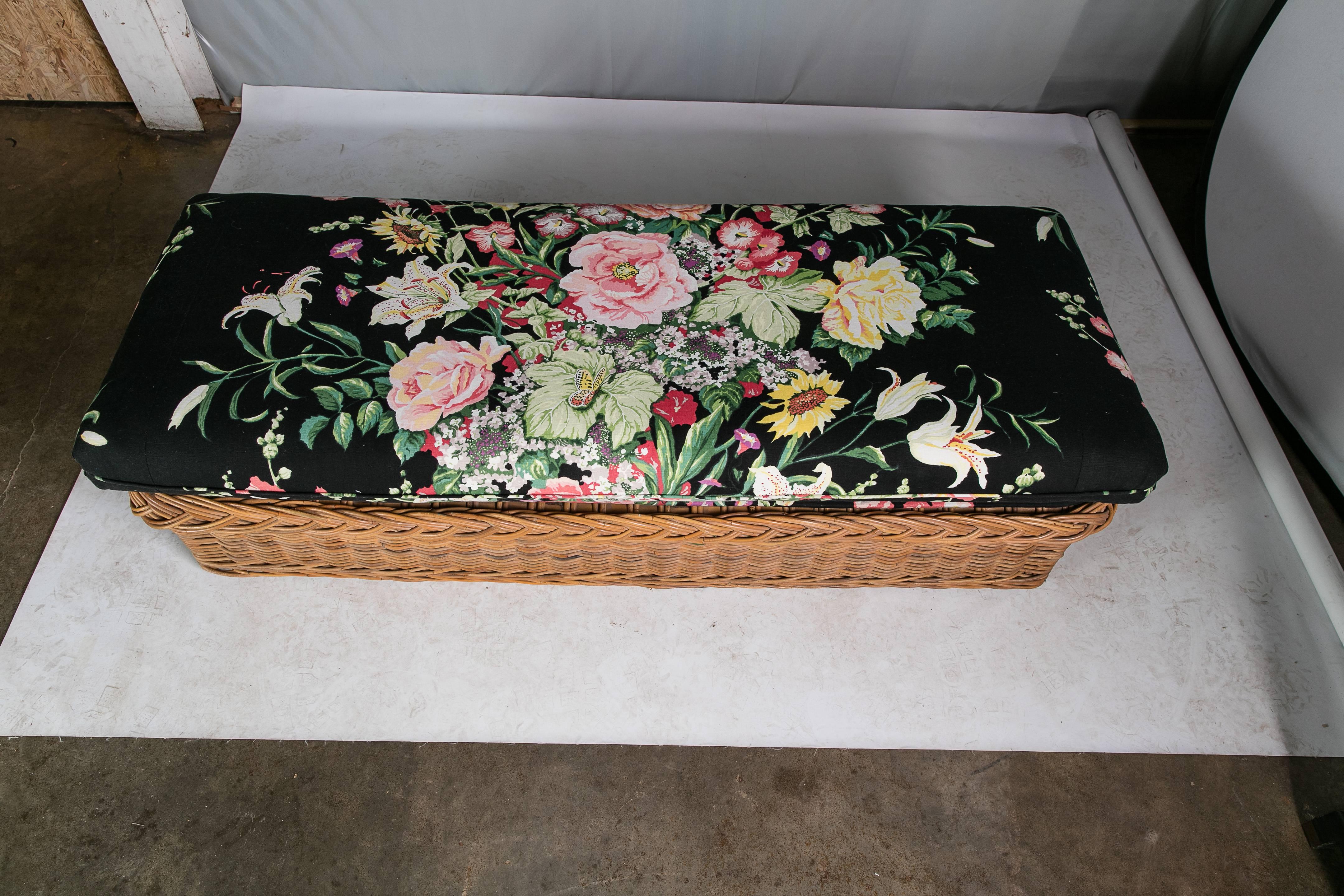 An Italian handwoven walnut-hued wicker bench with a loose rich black cotton cushion with a vibrant floral print. The cushion has a sturdy cotton wrapped foam interior. The deck under the cushion is upholstered in the same fabric as the cushion.