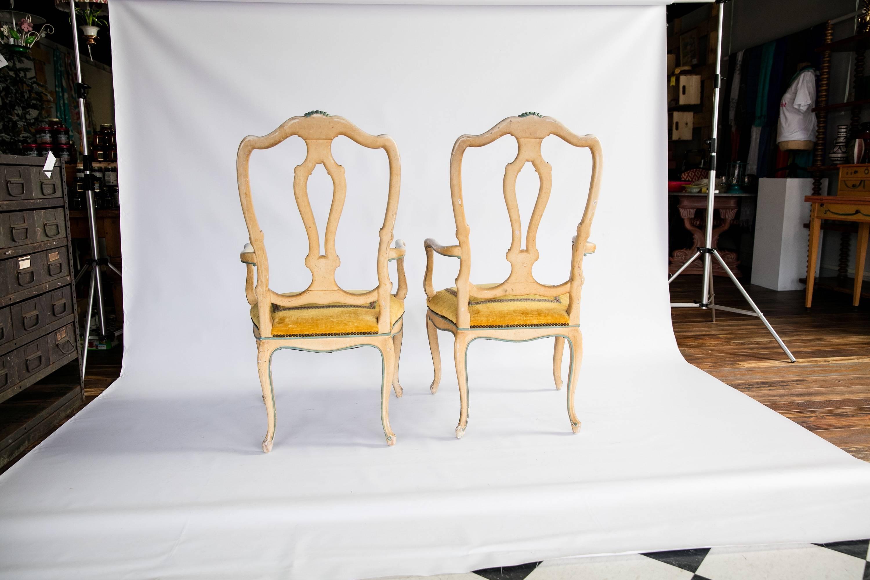 A pair of Venetian style arm chairs from Baker Furniture with original yellow velvet seats trimmed in pink-blue-and-green tape and brass nailheads. The chair frames are painted a creamy white and beige with blue-gray highlights and shell motifs.