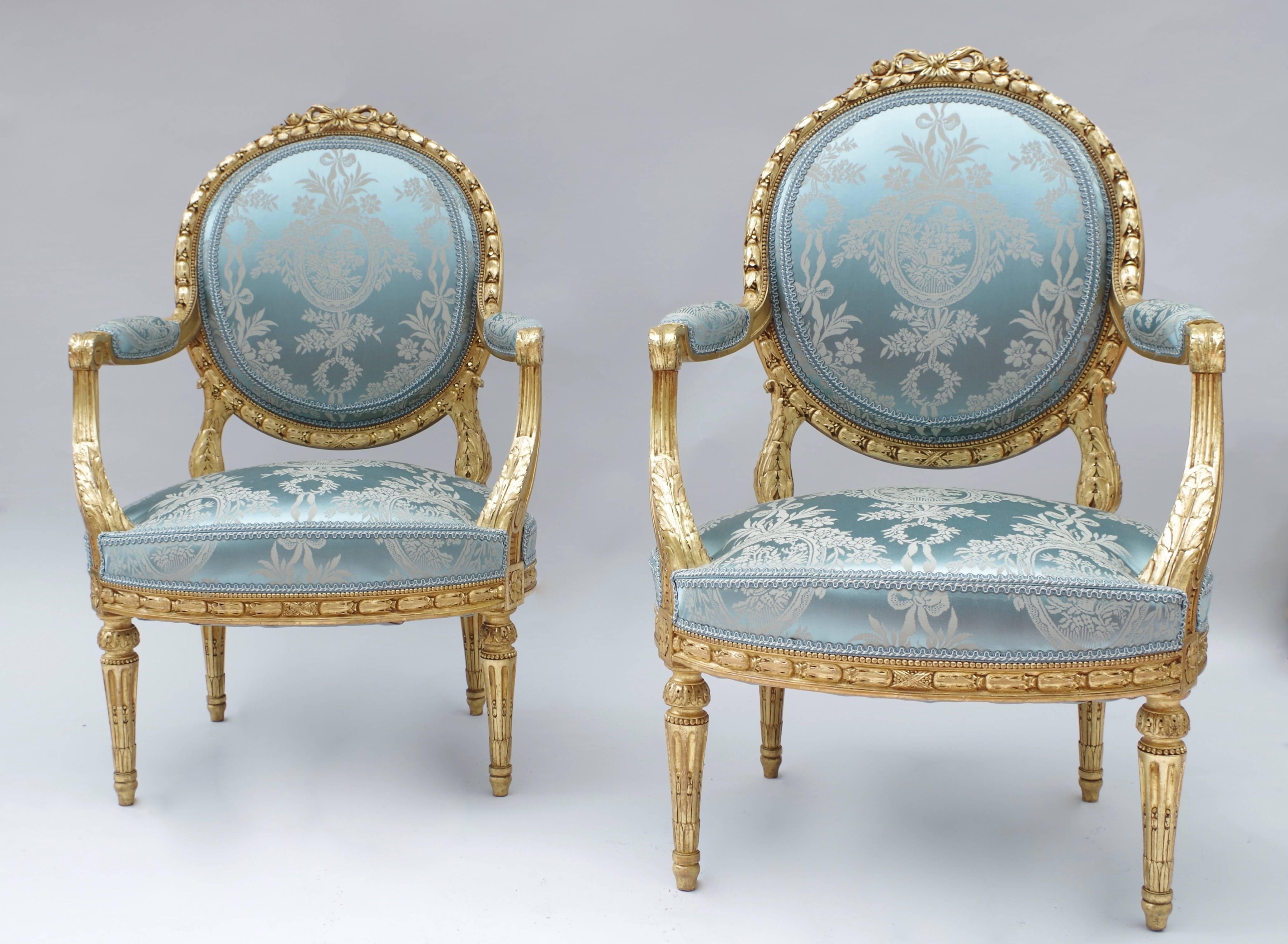 Pair of Louis XVI style giltwood armchairs with a flat backrest in a medallion shape. They rest on four legs with fluted shafts. The giltwood is carved with decoration of ribbons, garlands of laurel wreaths and acanthus leaves, in the classical