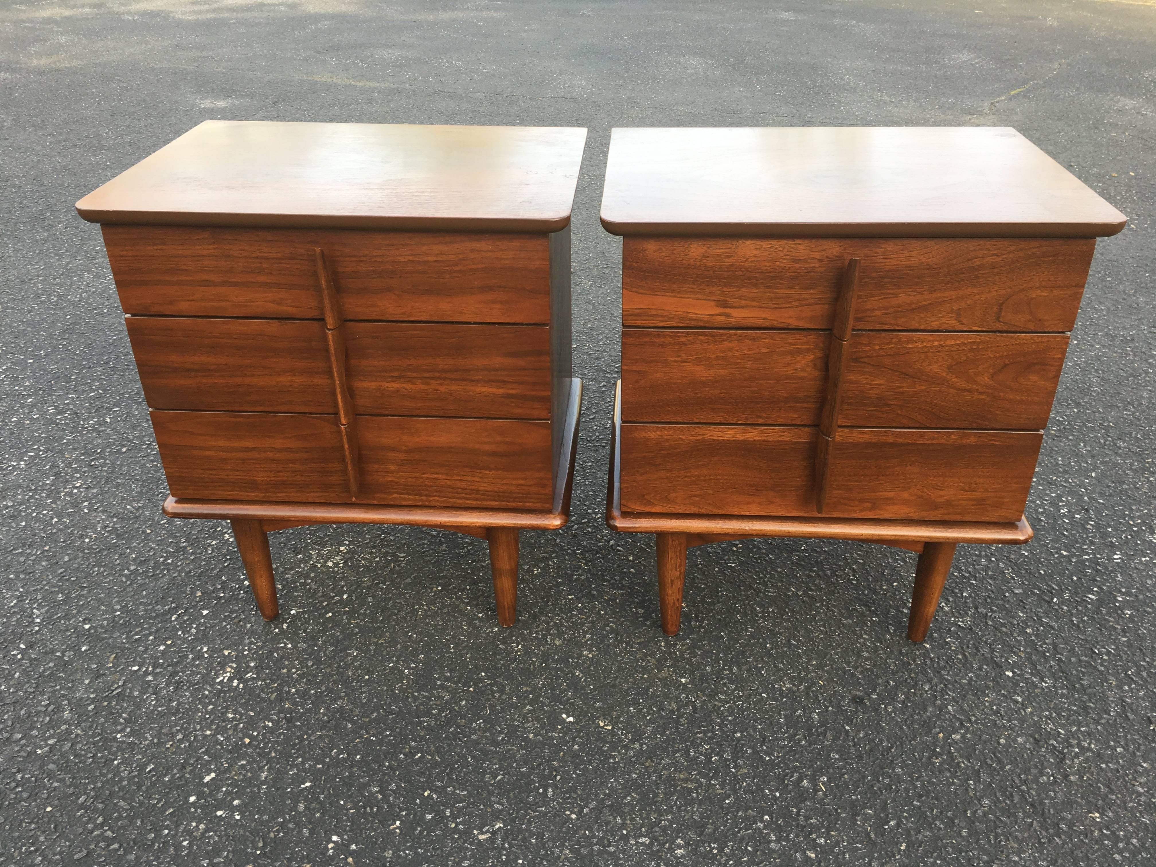 Pair of Mid-Century Modern nightstands. Sculptural handles adorn these Walnut beauties. They consist of three drawers each and have a composition banding below the wooden top. The small dot on the top right rear nightstand was wax and has been