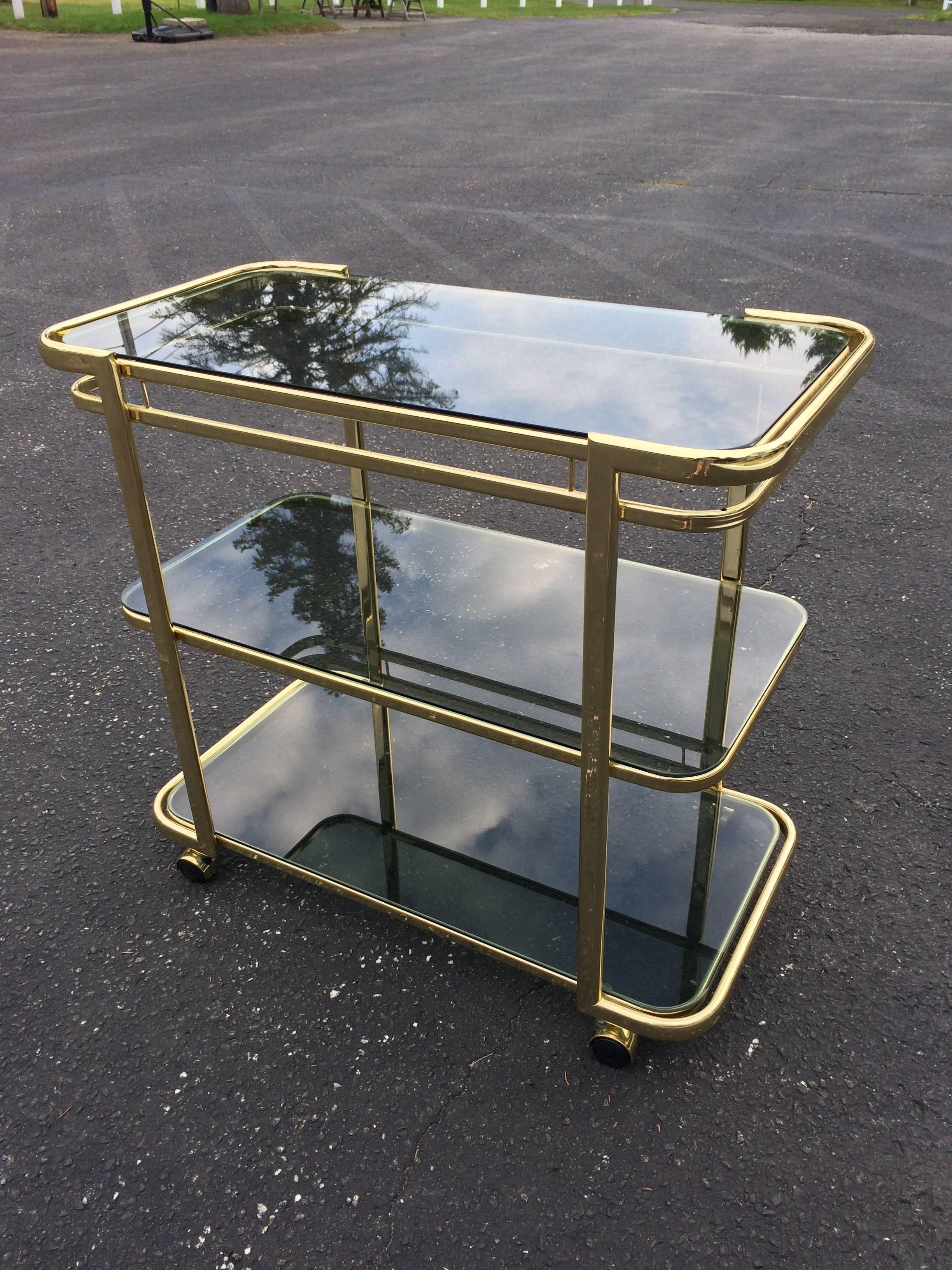 Brass and smoked glass bar cart. Sleek minimalist lines make up this beauty. The smoked Glass gives it an added sexiness and contrasts well with the polished brass. Three shelves allow for ample serving space for glasses and bottles or dessert and
