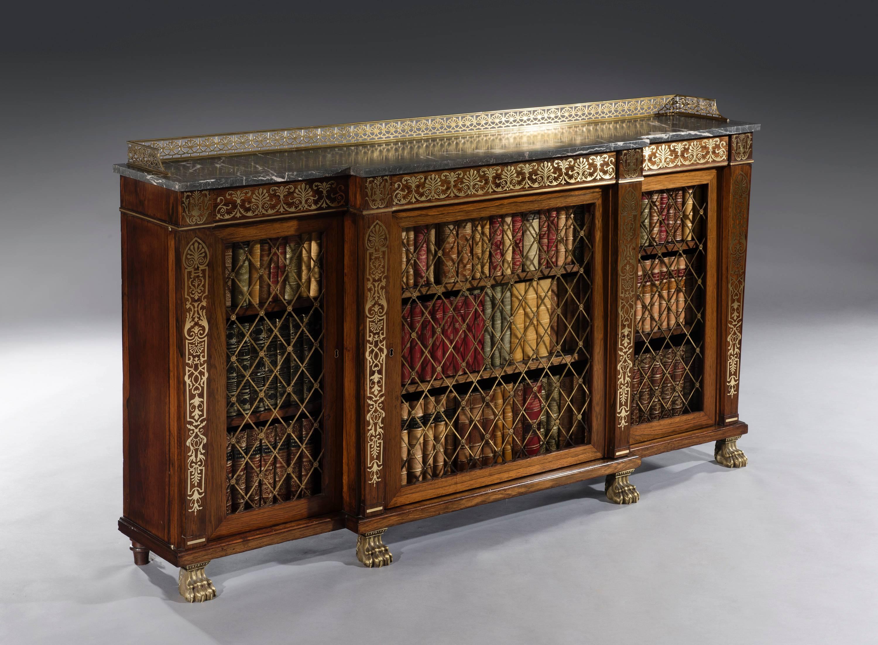 The breakfront bookcase retains its George Oakley trademark anthemion decorated open fret brass gallery and St. Anne marble-top above a brass strung inlaid frieze with ornate inlaid blocks above four elaborate brass inlaid tapering columns. The