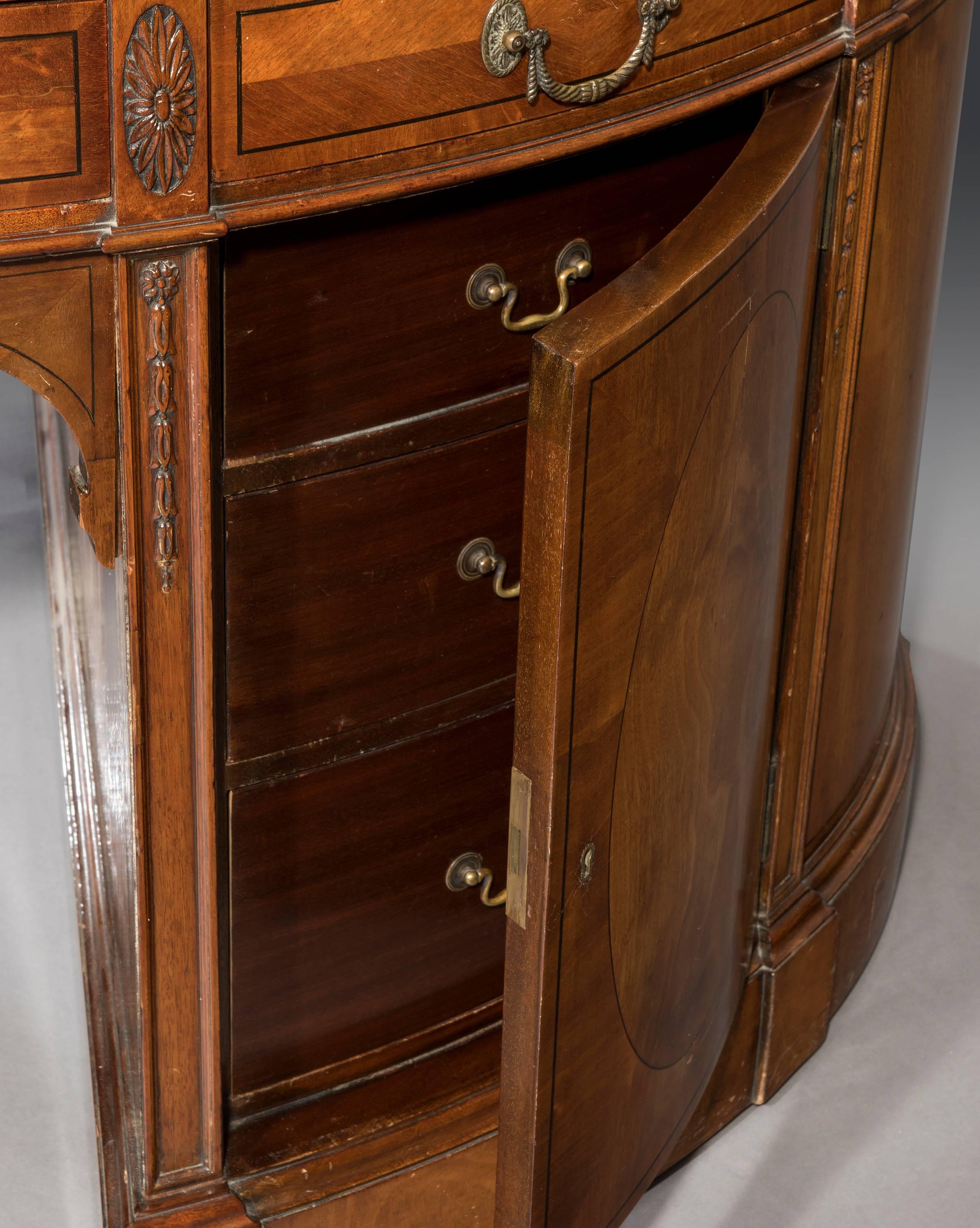 The rare oval-shaped breakfront form of this mahogany partners pedestal desk retains a brown tooled leather top above ebony lined frieze drawers flanked by carved patarae. The contrasting bookmatched veneers and ebony stringing to the drawer fronts