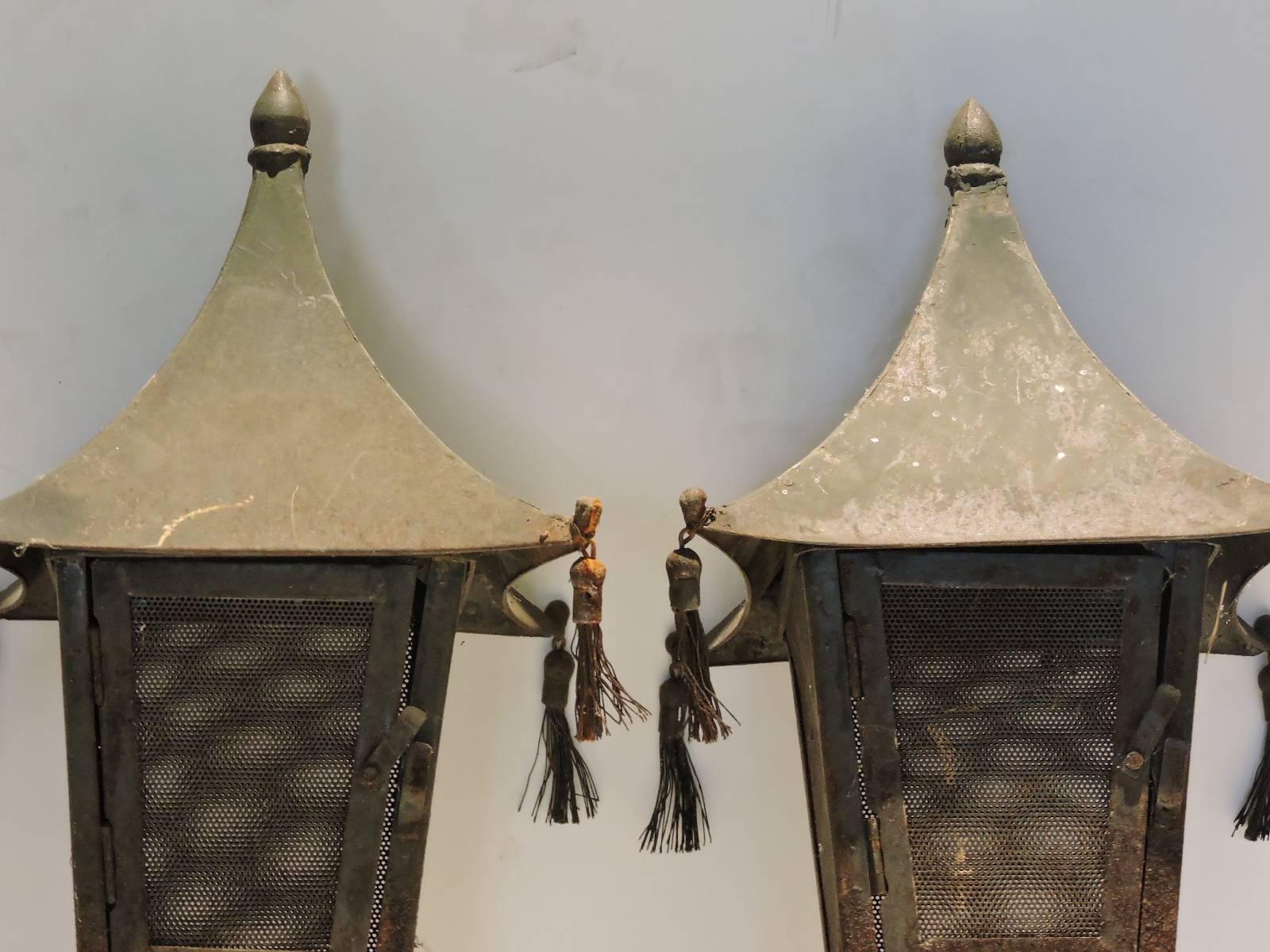 A good looking pair of tin metal pagoda form tasseled candle lanterns in original old worn aged gray to slightly green painted surface with areas of rust from being exposed to the outside elements for many years.