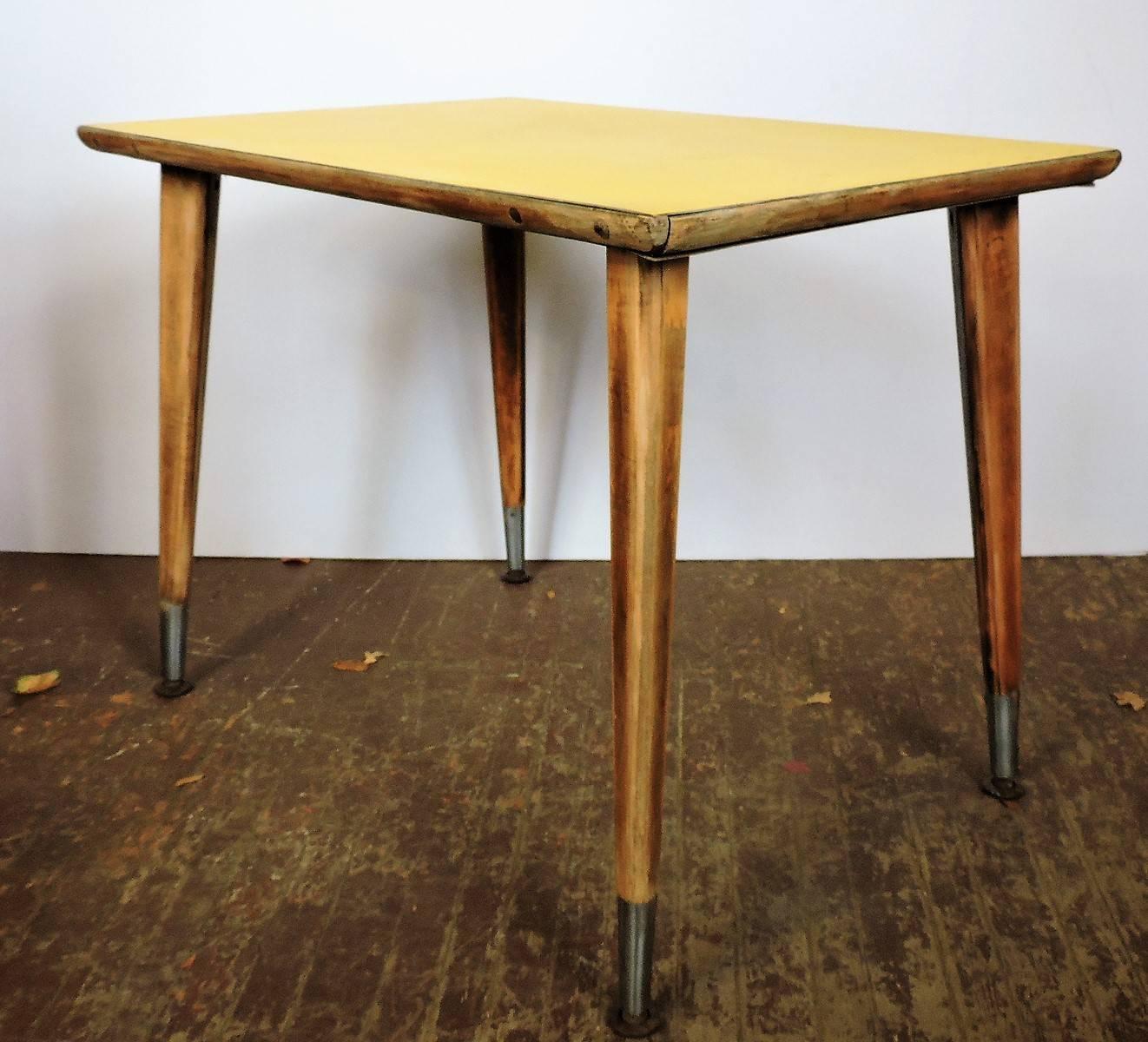 In the style of Jean Prouve - a Mid-Century Industrial table with beautifully angled splayed legs ending in long aluminum foot sleeves with swiveling foot caps. The top in original aged yellow laminate surrounded by pegged convex molding. Beautiful