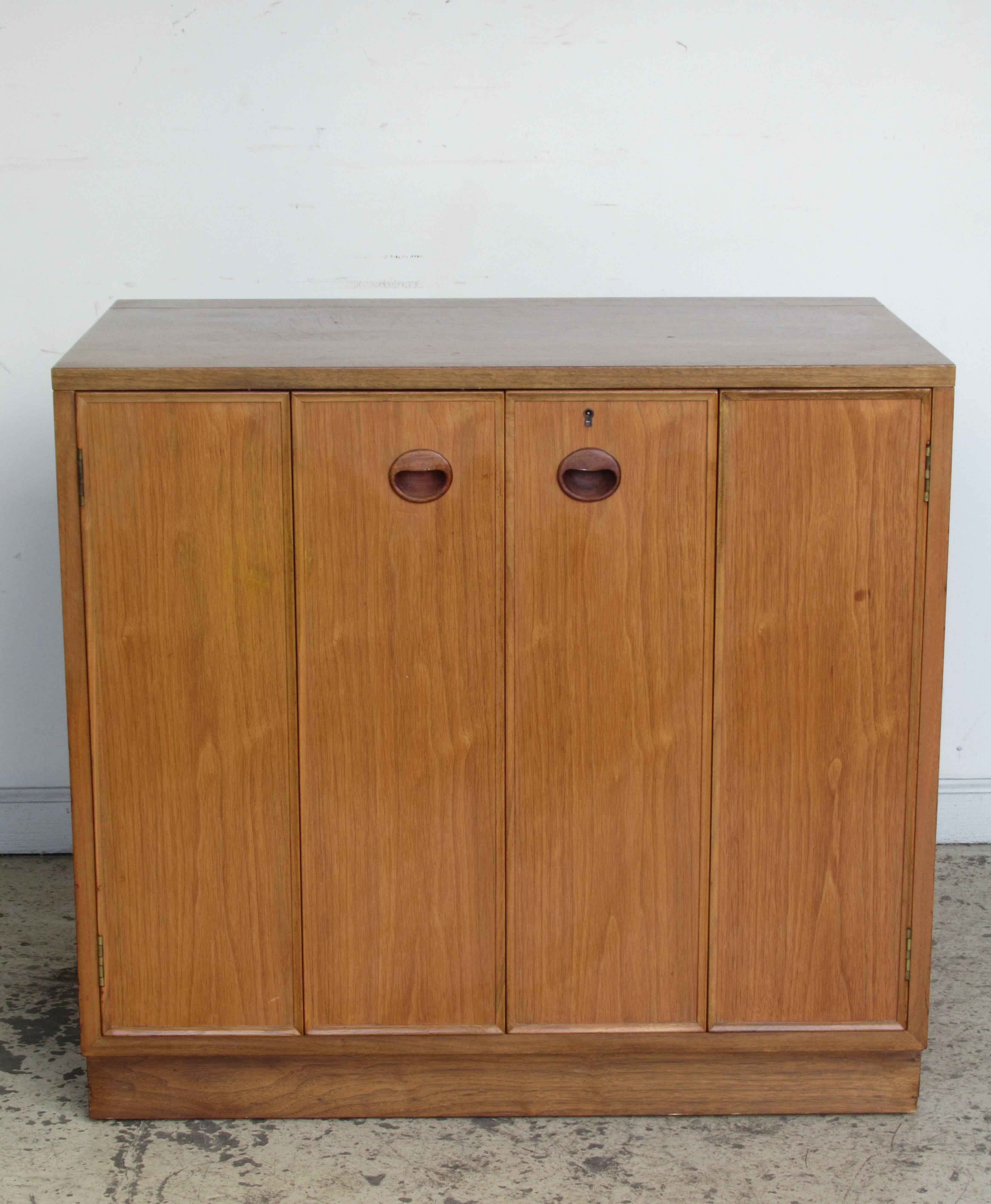 A walnut dry bar cabinet by Edward Wormley for Dunbar. The cabinet with a brass hinged lift top / two-fold accordion doors / fitted interior of 1 -2 white laminate top shelves / two sliding drawers and two open compartments. Great compact size that