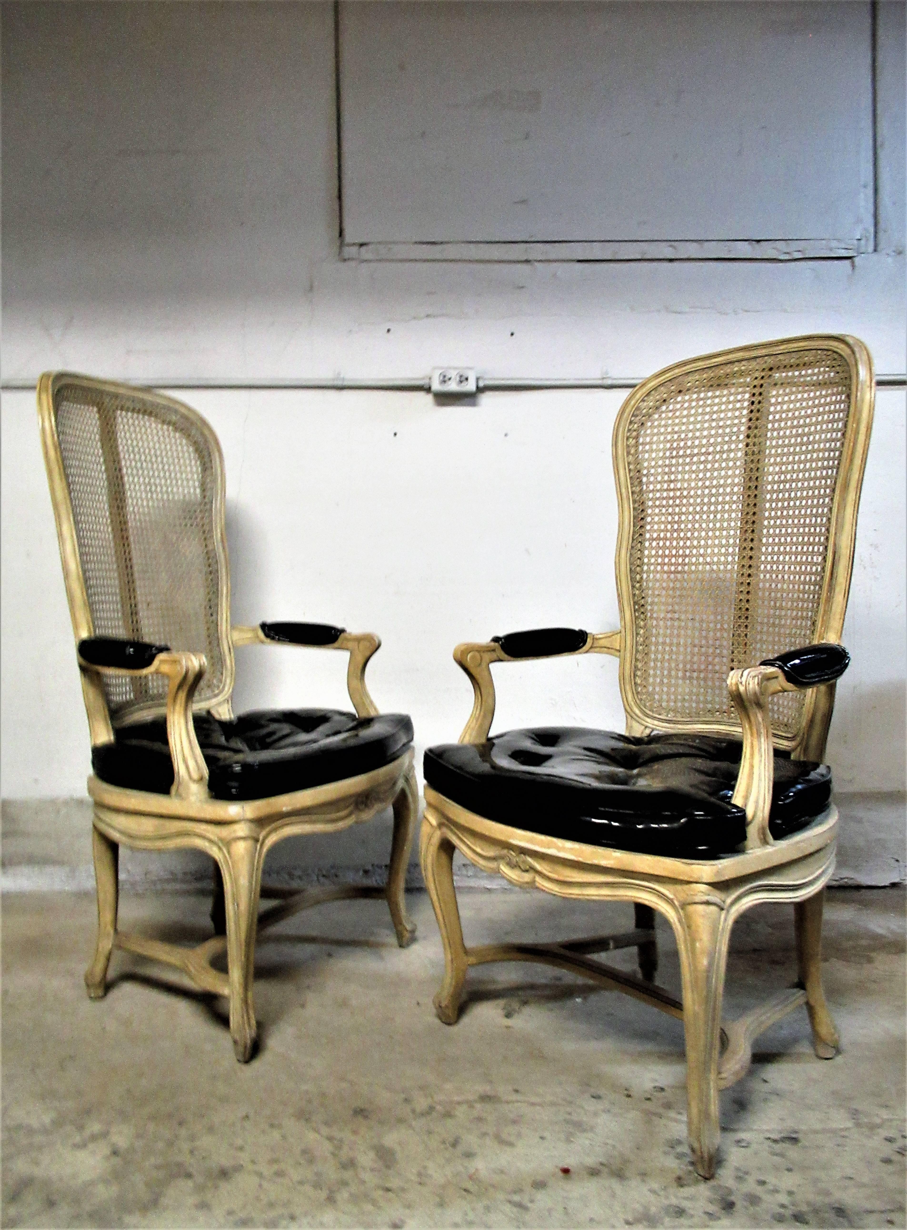 Pair of original pale cream painted Hollywood Regency tall cane back chairs in the Louis XV style with seat cushions and armrests in a high gloss black patent faux leather. Most likely Italian, circa 1960s. Good looking chairs.