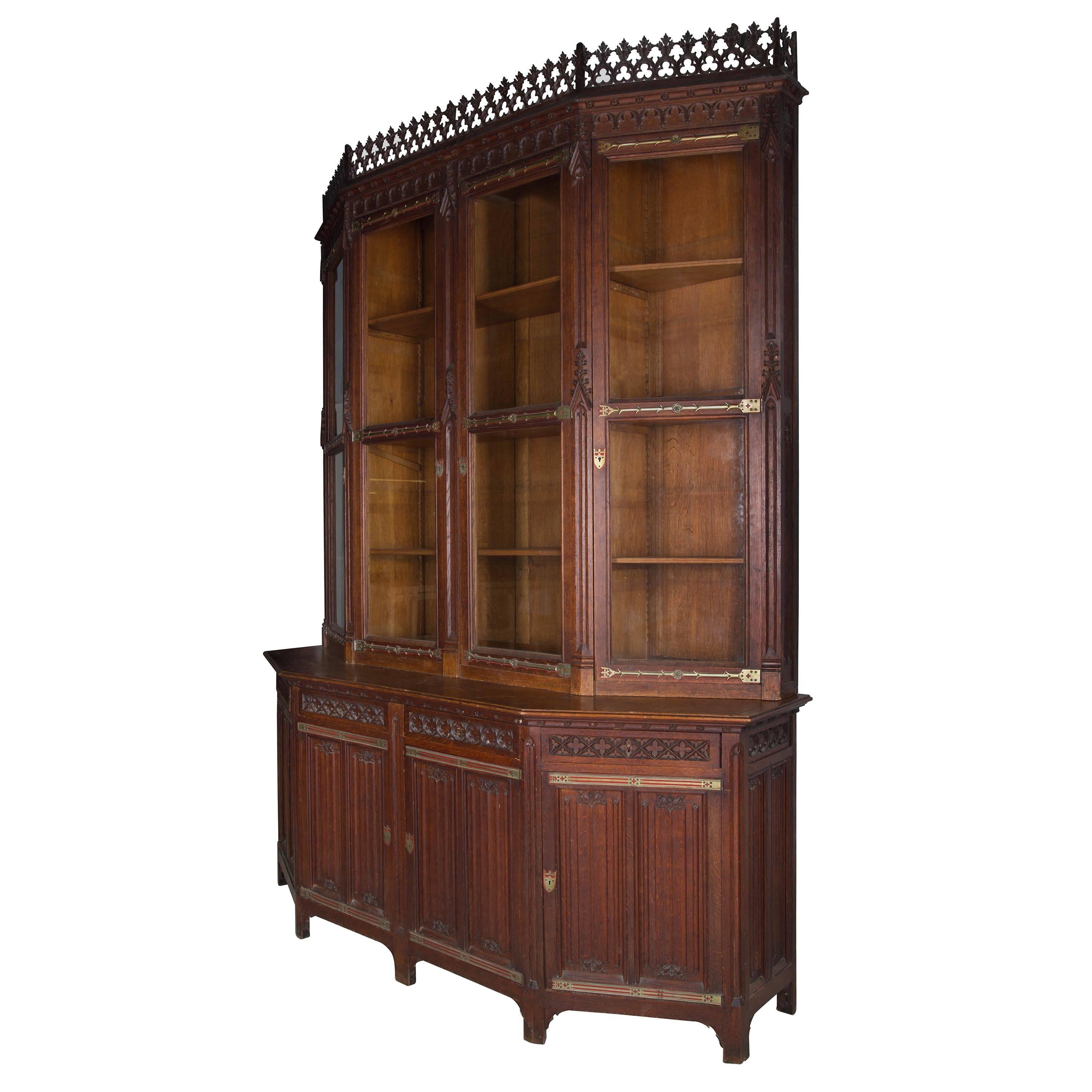 A Victorian carved oak Gothic Revival library bookcase, possibly after a design by Sir George Gilbert Scott, of canted form, the upper section fitted with four plain glazed doors above a base with four frieze drawers and blind cupboards below, the