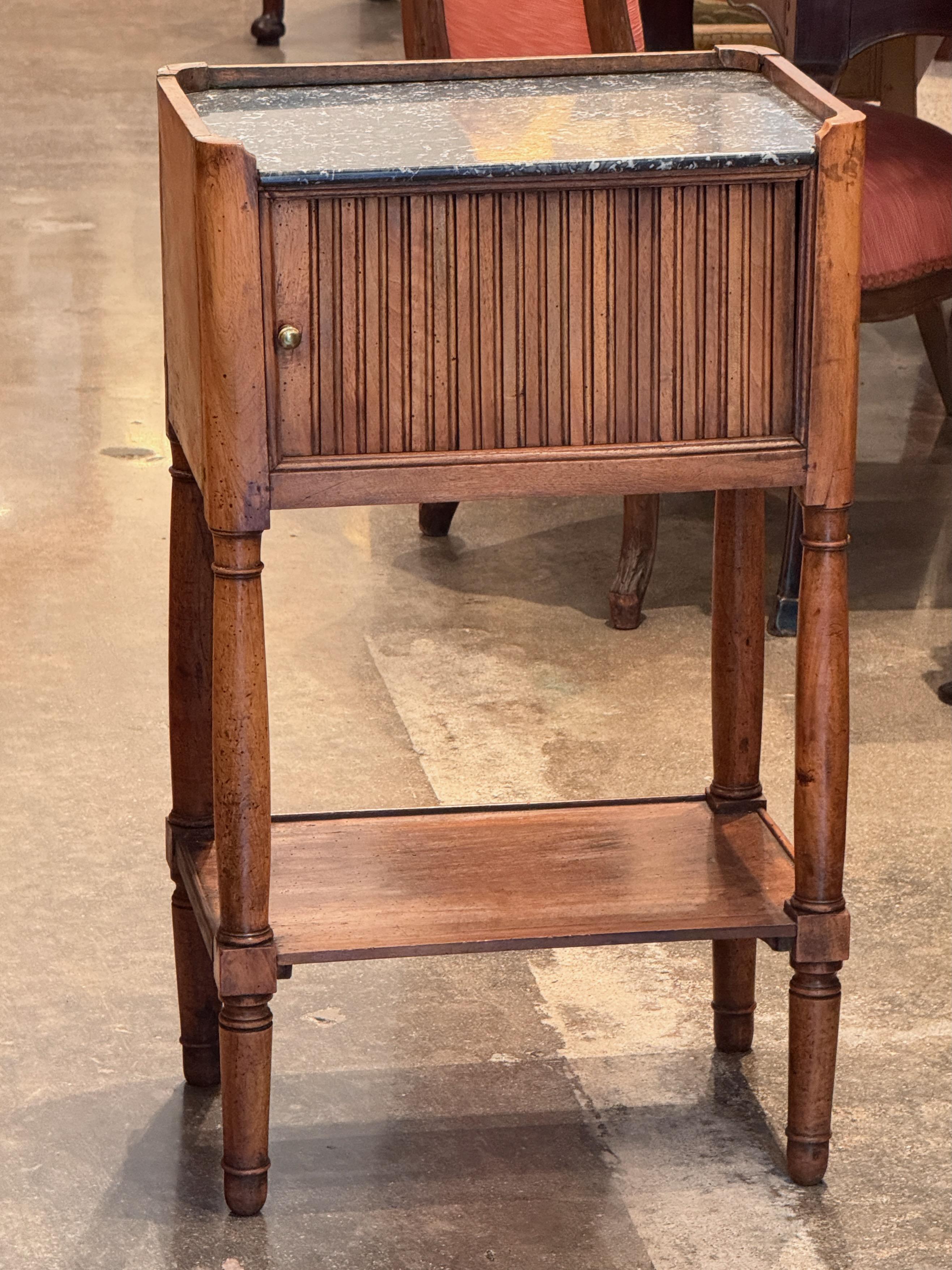 Narrow 19th century French side table in walnut, featuring a tambour-front storage compartment beneath a black marble top with raised gallery corners. Turned legs support a lower shelf, adding balance to the slender form. A practical and