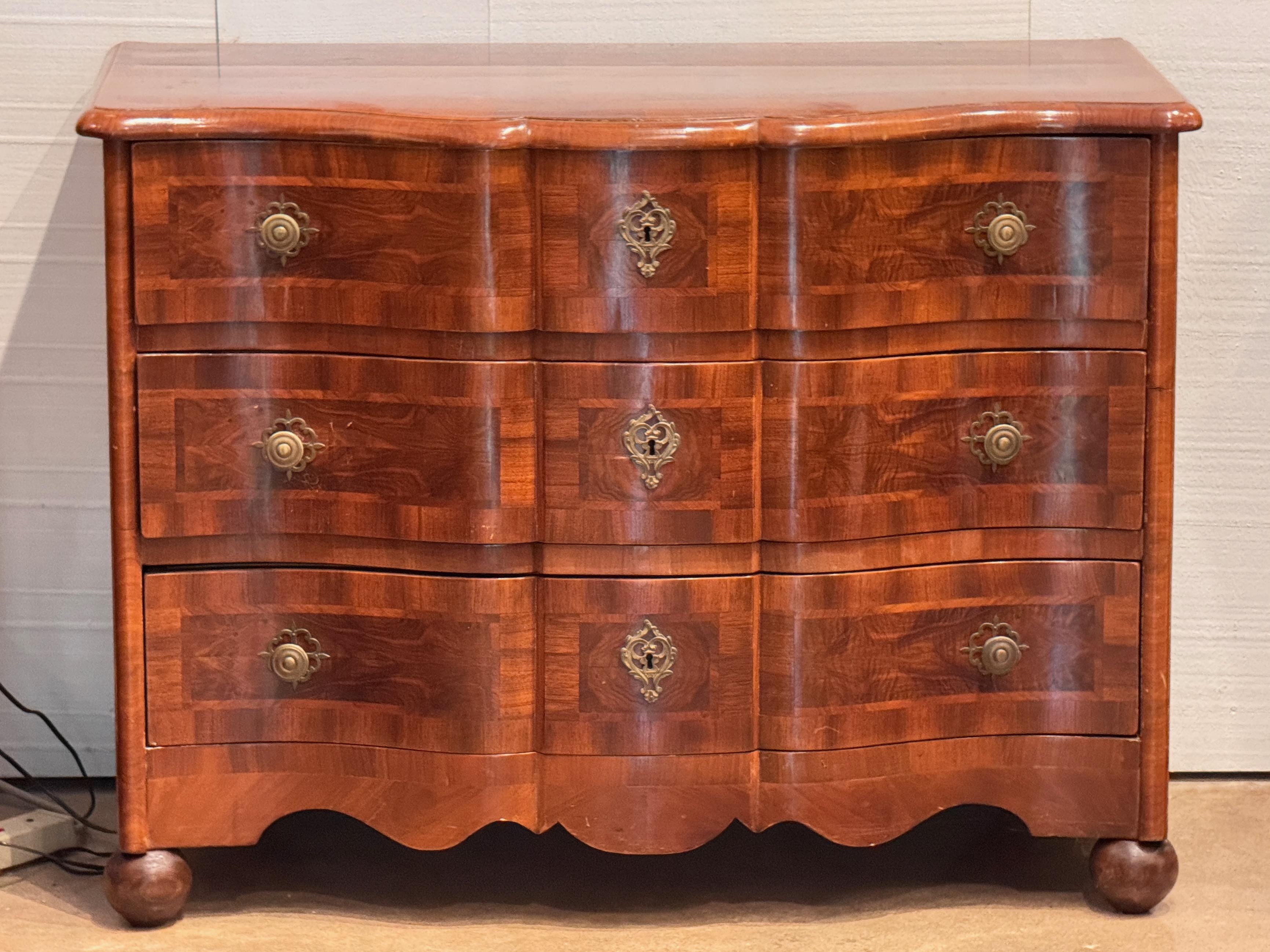 Three-drawer Continental chest with a serpentine front and shaped apron, featuring warm-toned inlaid veneers arranged in a geometric parquetry pattern. Original brass hardware and deep dovetail joinery point to fine early craftsmanship. Stands on