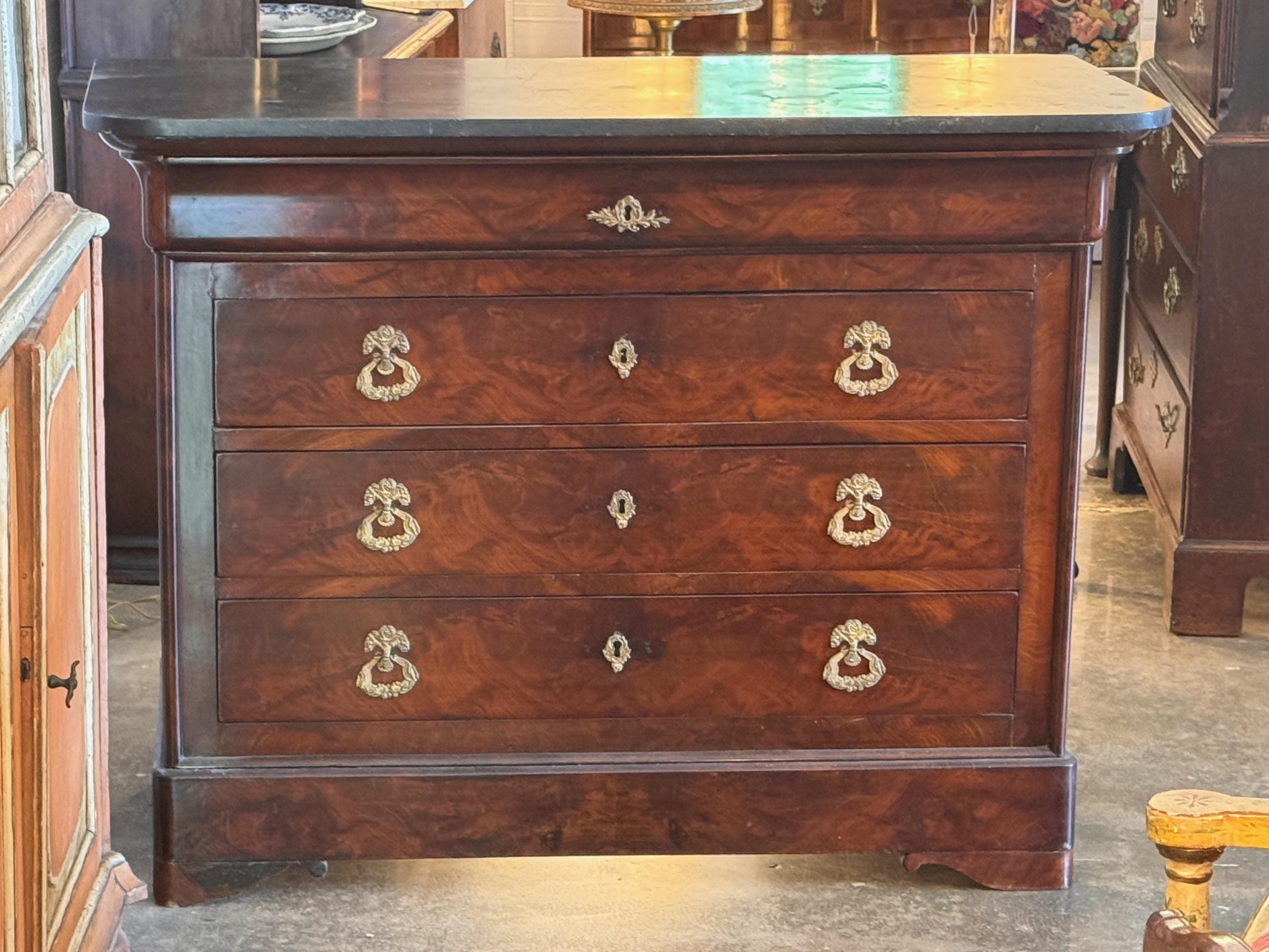 Louis Philippe period commode featuring a dark marble top over a case of matched veneer drawers with rounded corners and brass drop pulls. The bold grain and restrained design are characteristic of mid-19th century French style, offering both