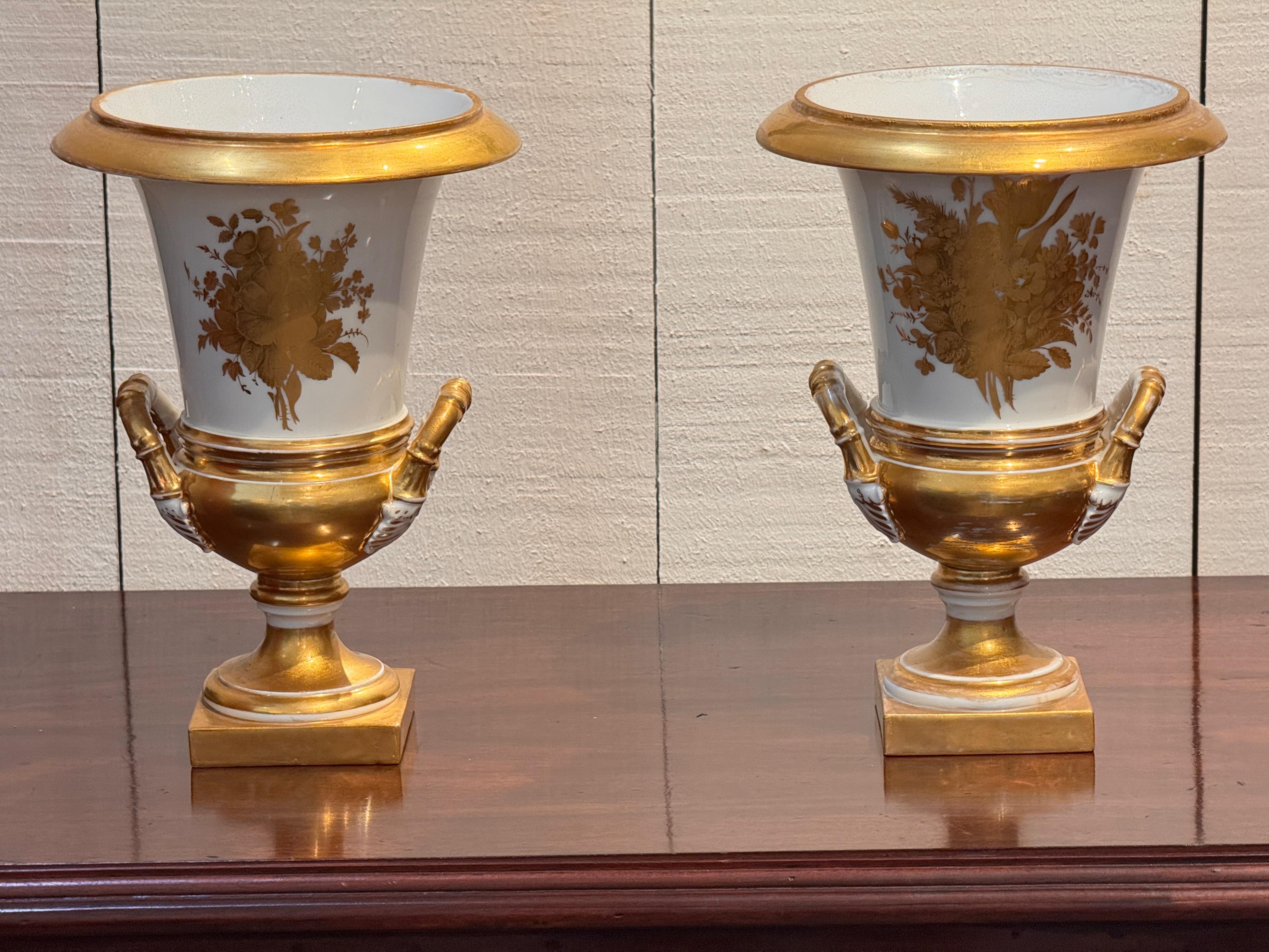 Coppia di urne in porcellana Old Paris del XIX secolo con manici dorati e basi quadrate con piedi. Ognuno di essi è decorato con spruzzi floreali dorati su un fondo bianco, mostrando la raffinata influenza neoclassica tipica delle porcellane