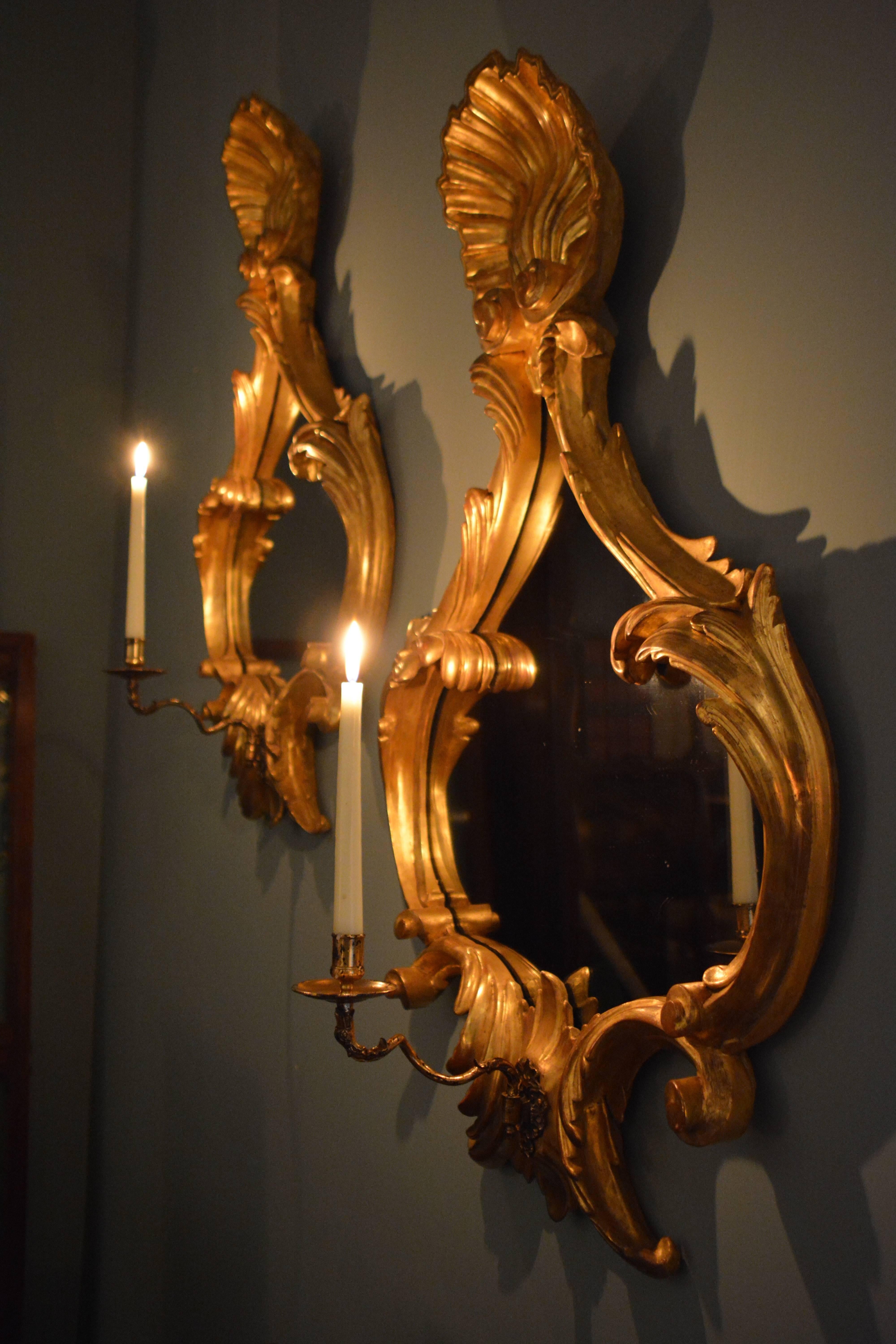 A rare pair of George II carved giltwood mirrors or girandoles, the scrolling frames surmounted by a carved shell, the base having a lacquered brass candle holder, English, circa 1730.