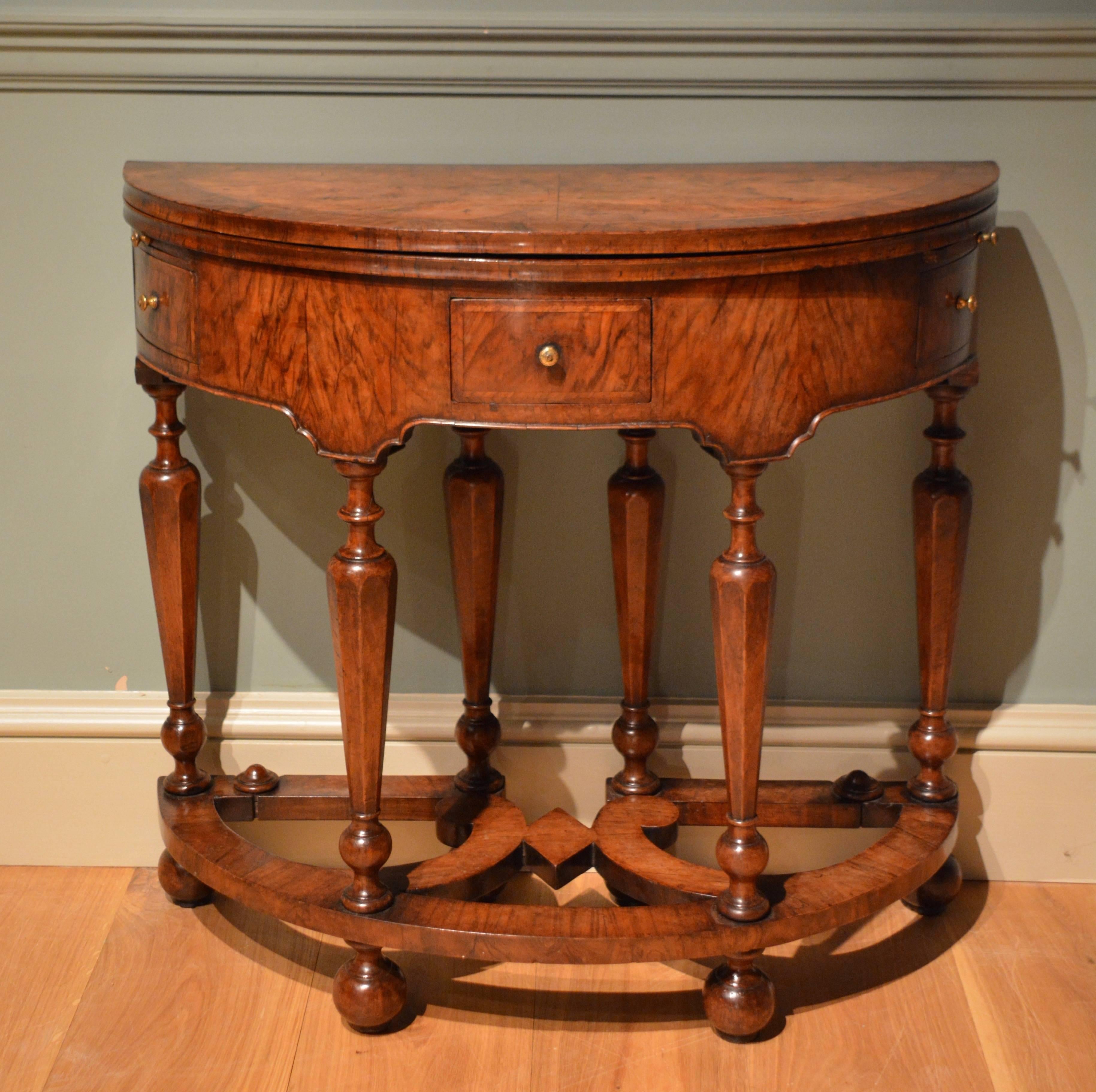 A Queen Anne veneered walnut demilune card table, the oval crossbanded and herringboned top opening to reveal a crossbanded velvet lined playing surface, the frieze containing two candle slides and three drawers, the whole standing on pillar legs