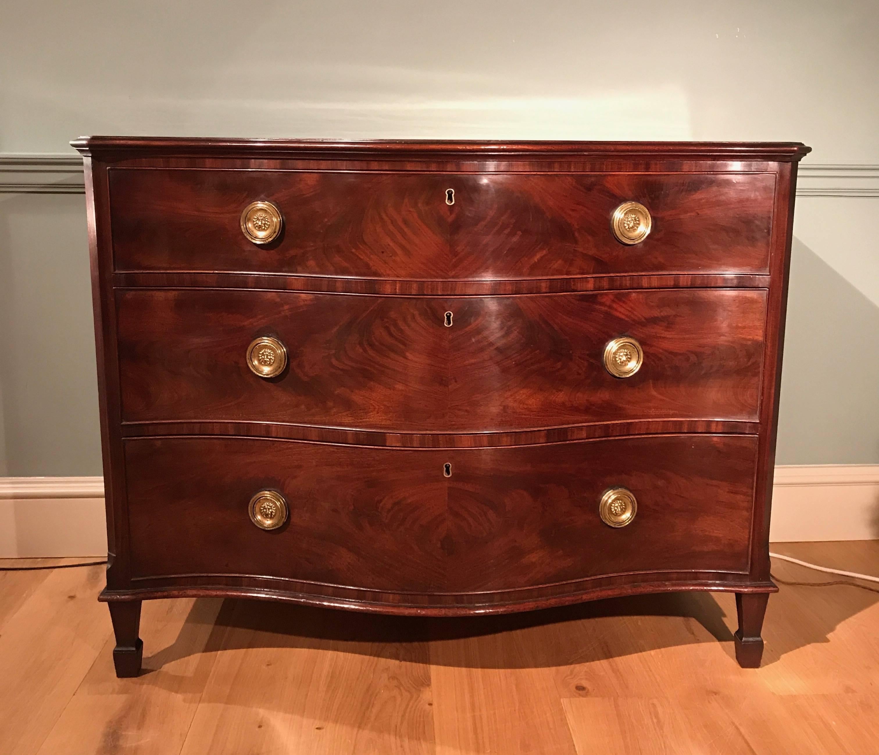A fine George III serpentine mahogany commode of excellent colour and patination, having three drawers with lion mask handles standing on toupe spade feet. English, circa 1790.