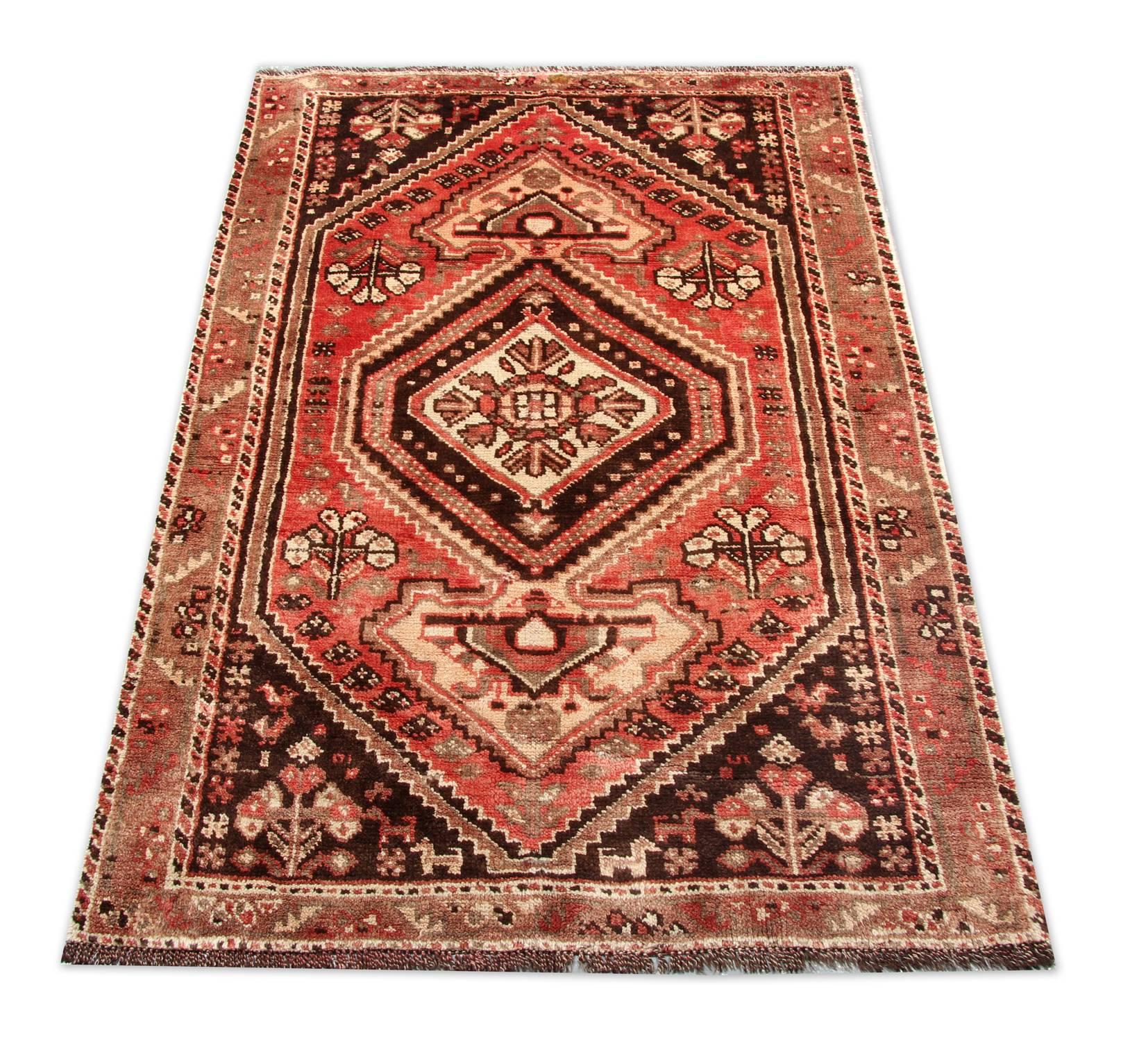 Esta fantástica alfombra de lana fue tejida a mano en Irán por maestros tejedores. Esta pieza presenta un intrincado diseño tribal simétrico tejido sobre un rico fondo rojo con acentos de color marrón beis, crema y naranja que componen el medallón