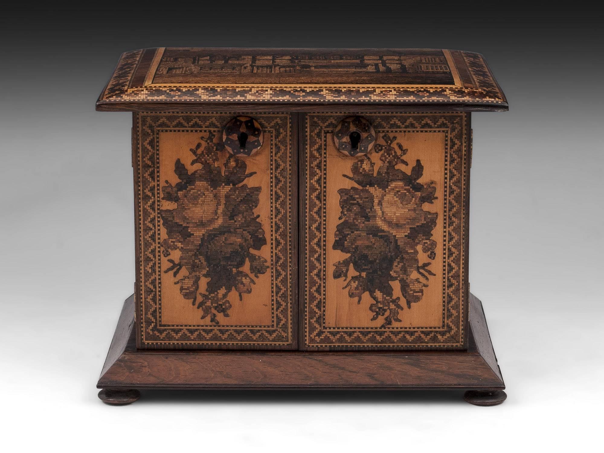 Tunbridge ware table cabinet veneered in Mahogany with floral inlays on the doors, a plinth base standing on four turned wooden feet. The top has a view of Shakespeare's birthplace. 

The interior of the tunbridge ware cabinet features four drawers
