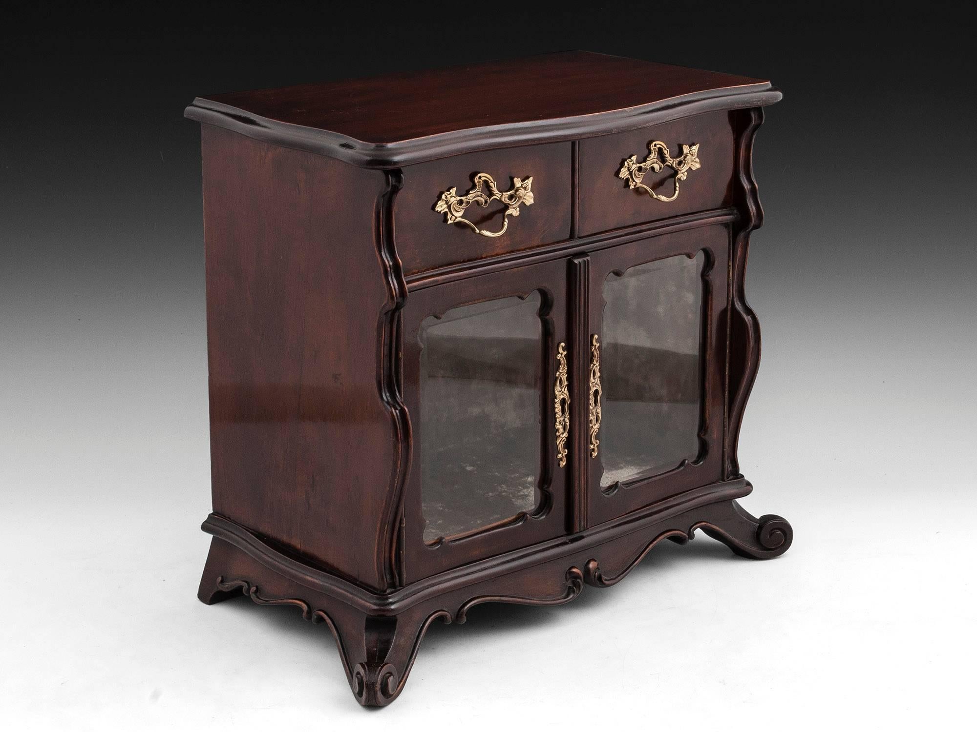 A fine quality walnut miniature display cabinet with scrolling carved feet and bevelled glass doors. 

Featuring two brass handled drawers and brass door escutcheons. 

This cabinet comes with a fully working lock and tasselled key. 