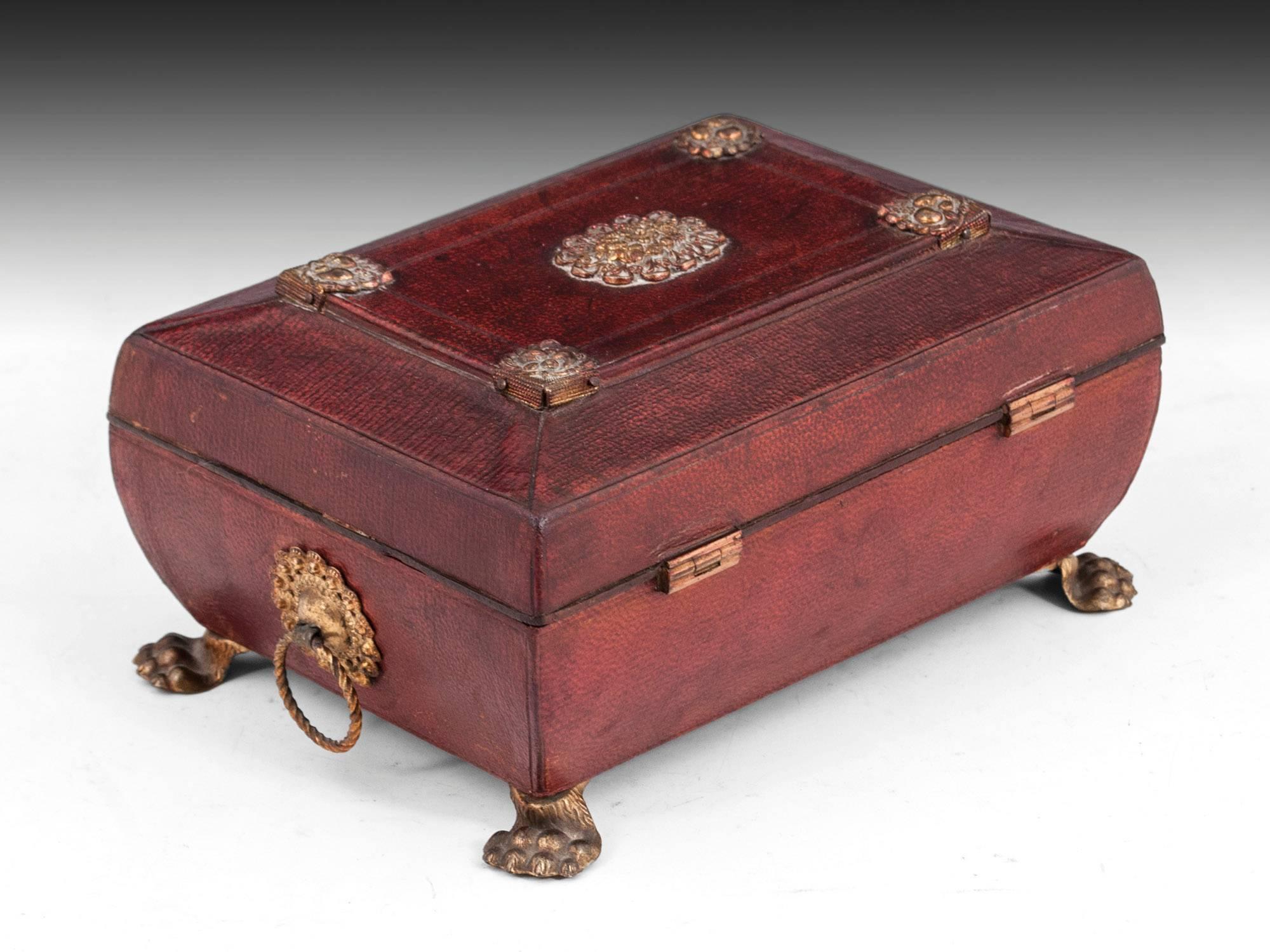 Miniature leather sewing box with a red leather exterior decorated with brass details, escutcheon, handles and pressed brass lions feet.

The interior is lined with blue paper and features a pincushion, needle book, toolpad, thimble tray and three