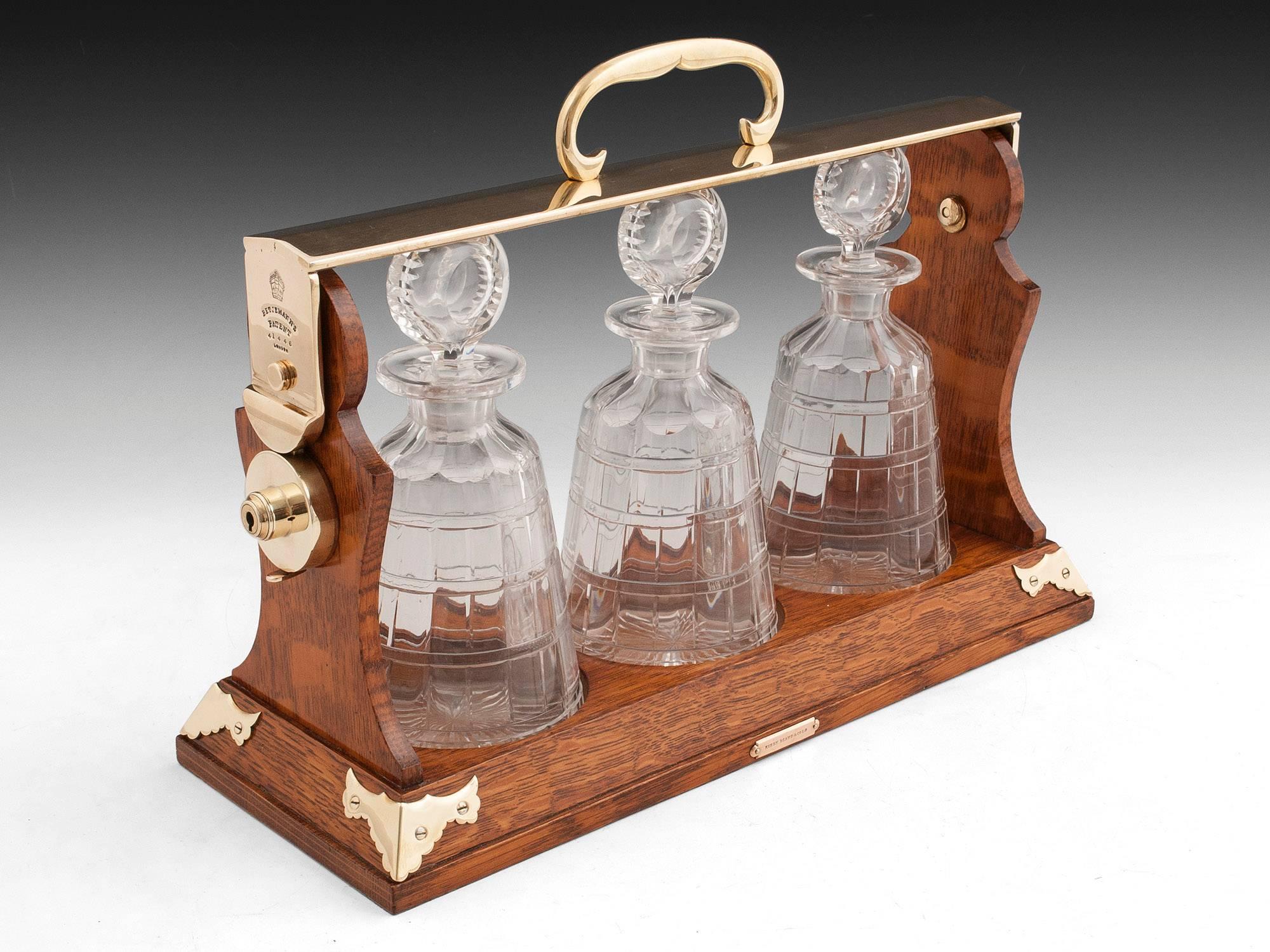 Oak Betjemann's Tantalus. Featuring ornate brass corner supports, carry handle and escutcheon.

The tantalus holds three unusual cut lead crystal decanters of cylindrical tapering form with cut-glass stoppers. The decanters can be removed once the