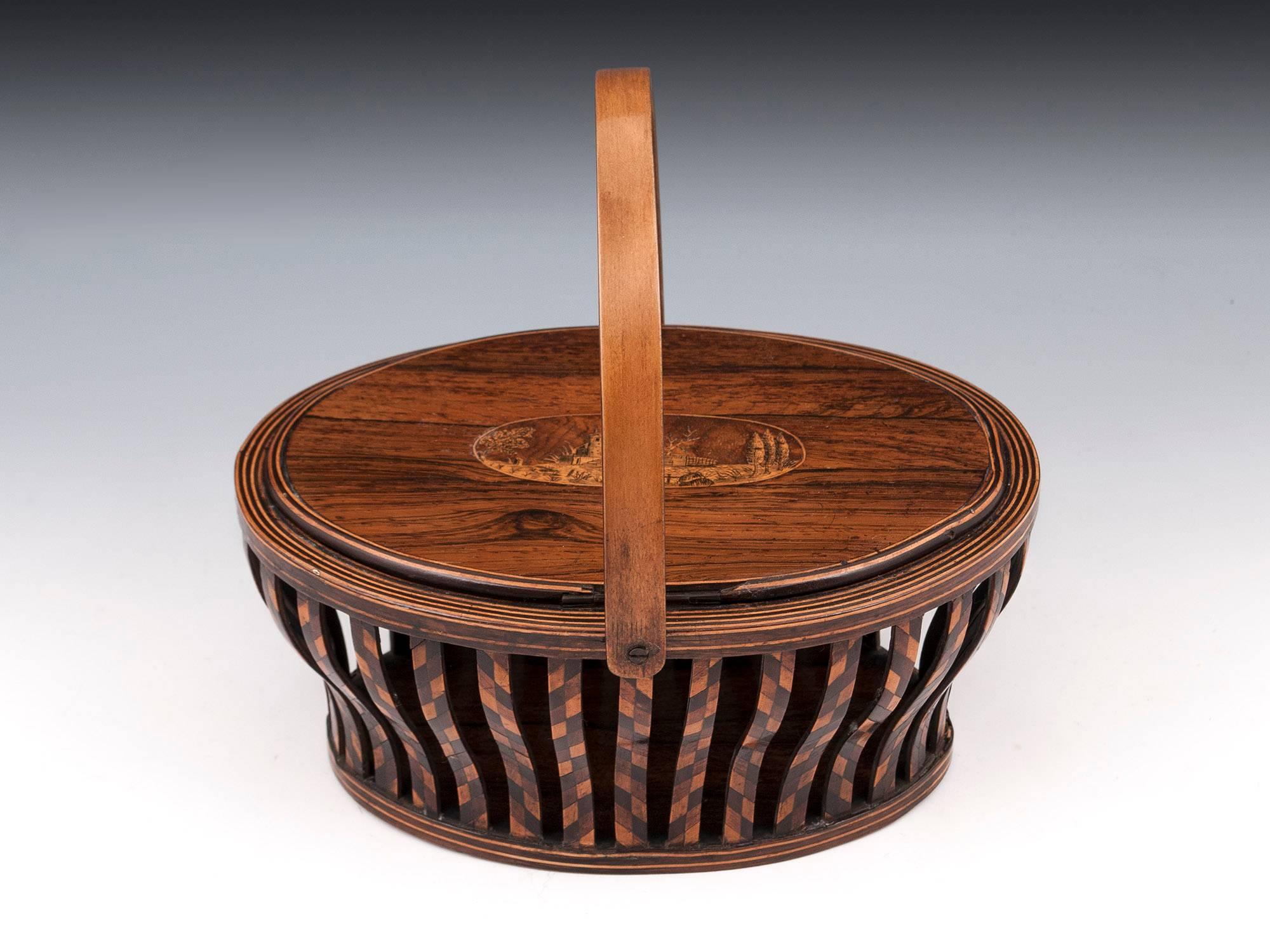 Antique sewing basket with shaped sycamore carry handle. On the top of the box is a naive penwork rural scene similar to Kilarney Ware. Each shaped side supports is veneered with a chequered design of Mahogany, Sycamore and Yew. The hinged lid has a