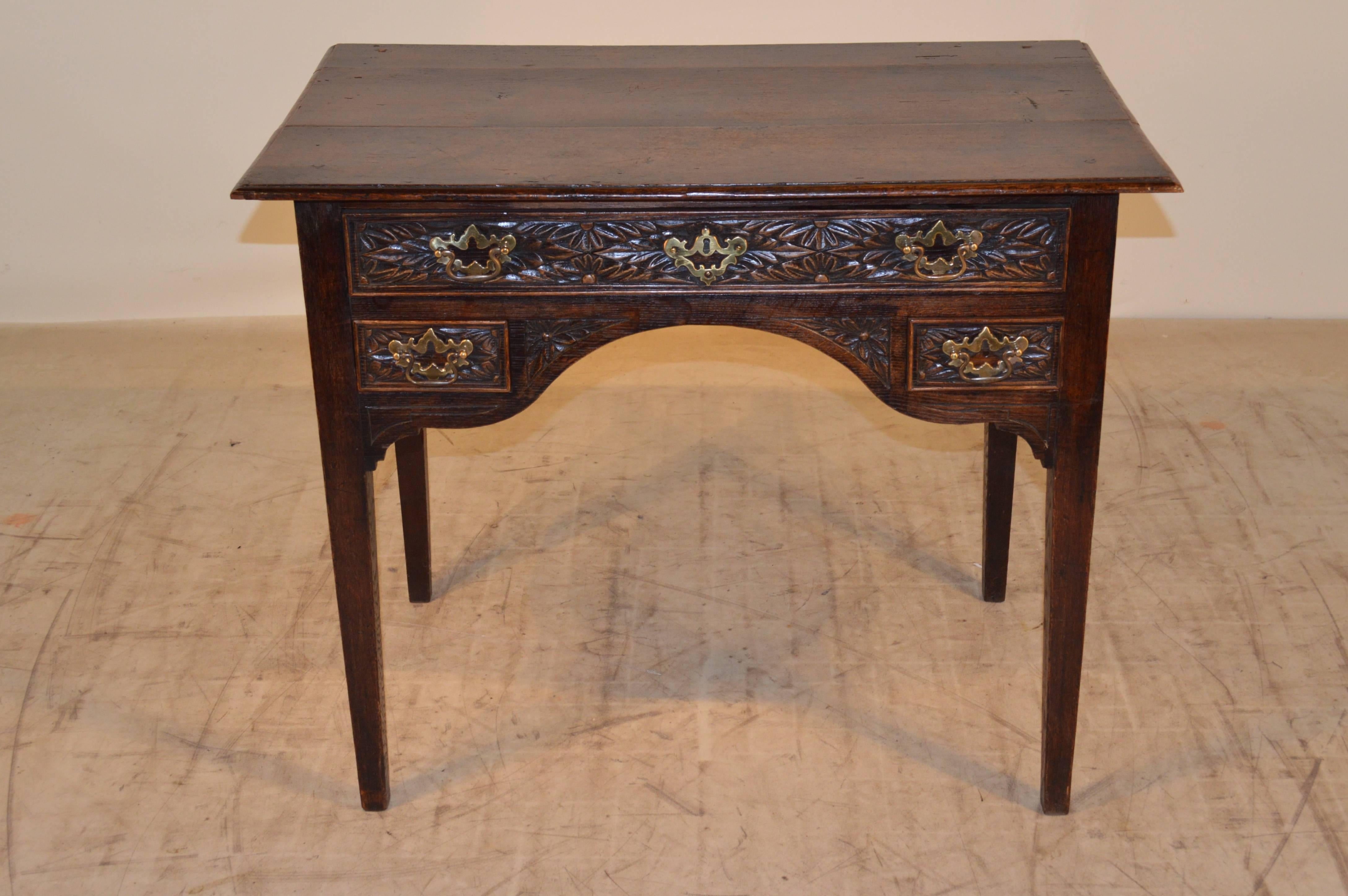 18th Century English oak lowboy with a plank top which has a beveled edge. The case has a shaped apron with three drawers in the front, all with carved decoration. The piece is supported on hand tapered legs. There is a slight nibble on the front
