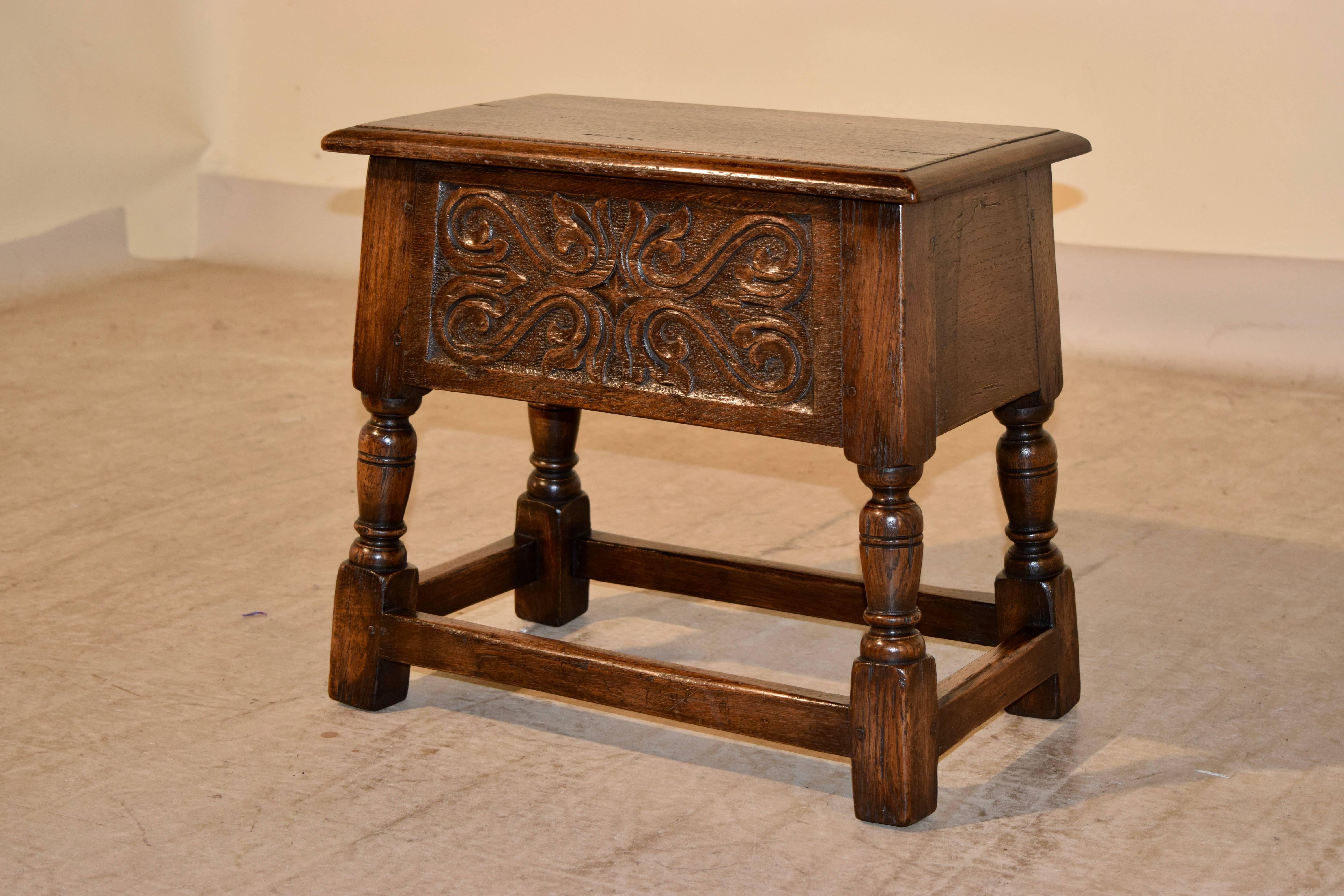 19th century oak lift-top stool from England. The top is beveled around the edge and opens to reveal storage. The front panel is hand-carved decorated, and is supported on hand-turned legs joined by stretchers. The top measures 19.88 x 12.