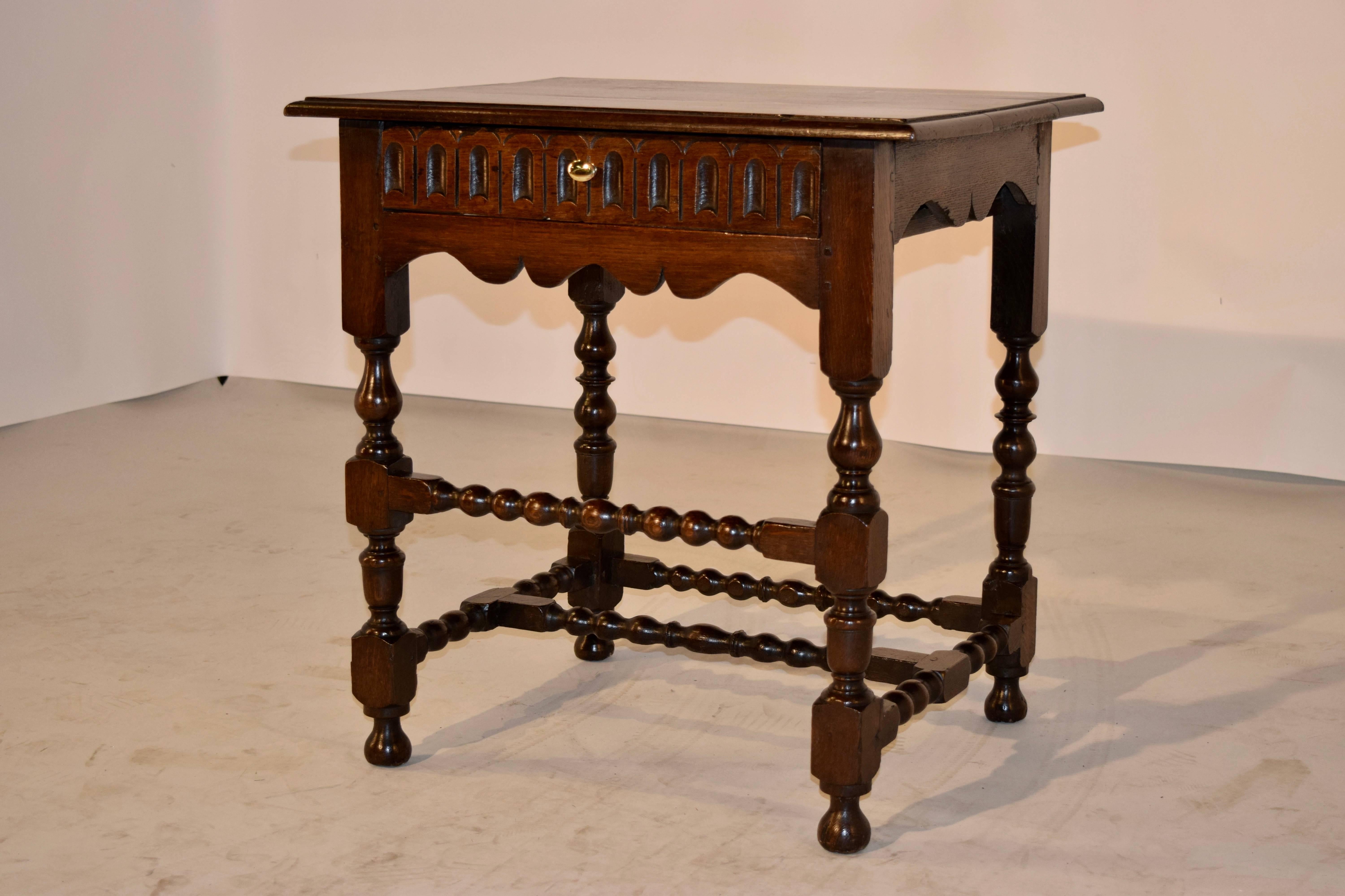 19th Century English oak side table with a beveled edge around the top following down to a single drawer with a carved drawer front and a scalloped apron. The legs are hand-turned and have complimenting turned stretchers and it is raised on turned