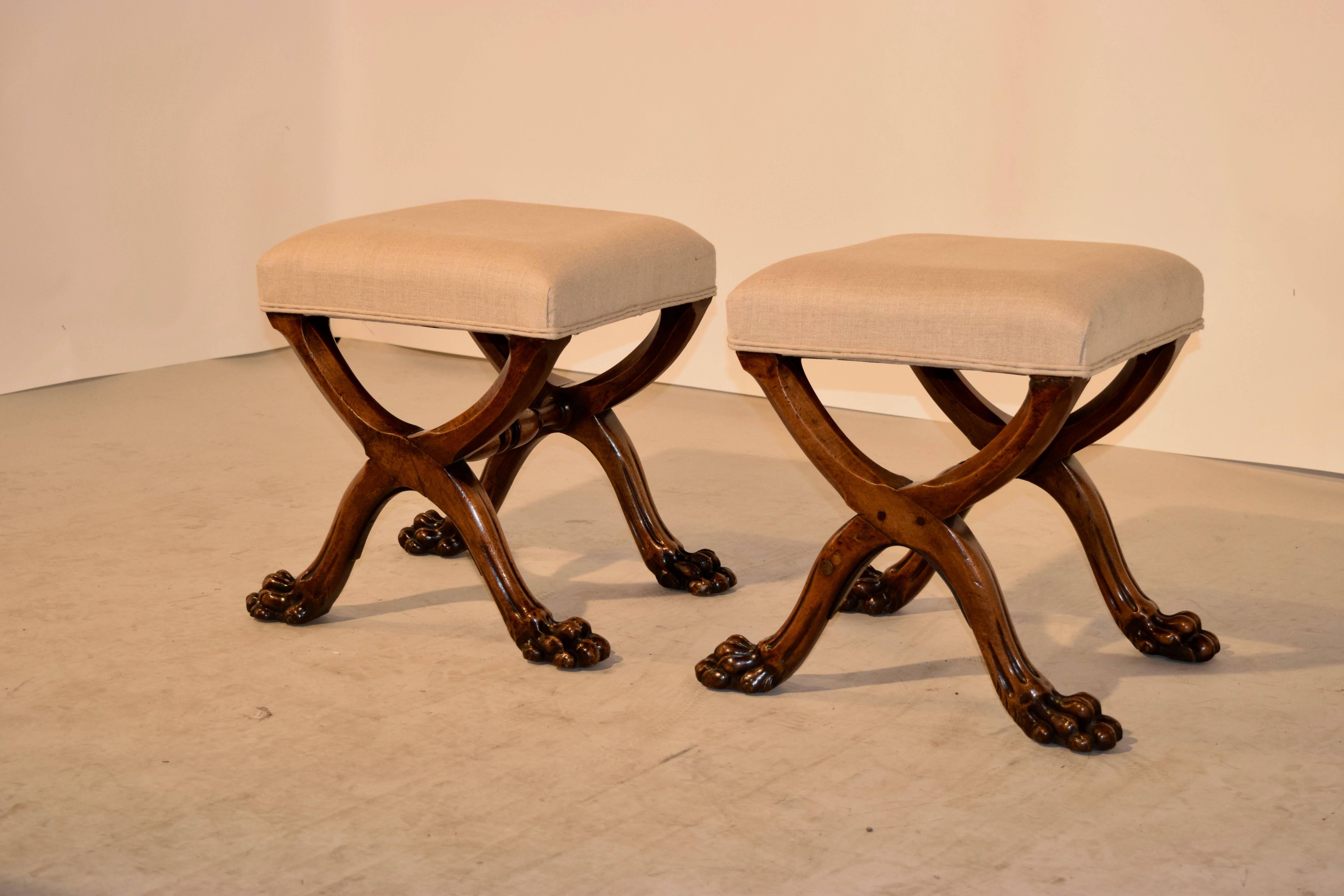 19th century pair of English stools with newly upholstered tops in linen. The bases are made from mahogany, as are the X-style legs, and joined by hand-turned stretchers ending in hand-carved paw feet. Newly upholstered in linen, old repairs and