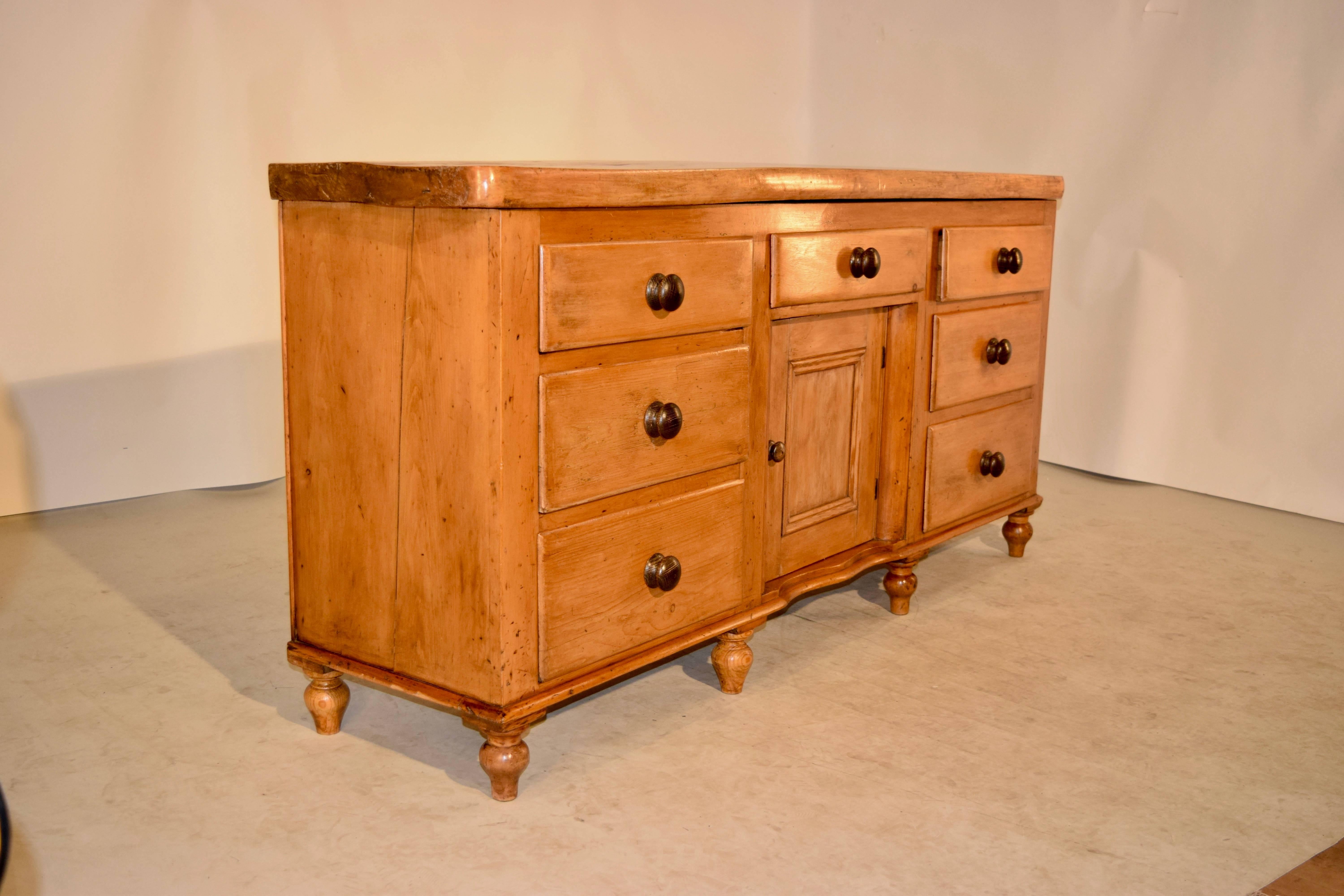 19th century English dresser with a thick single plank top made from sycamore, following down to a simple case with two banks of drawers flanking a single central drawer over a central door. The bottom is scalloped and the piece is resting upon