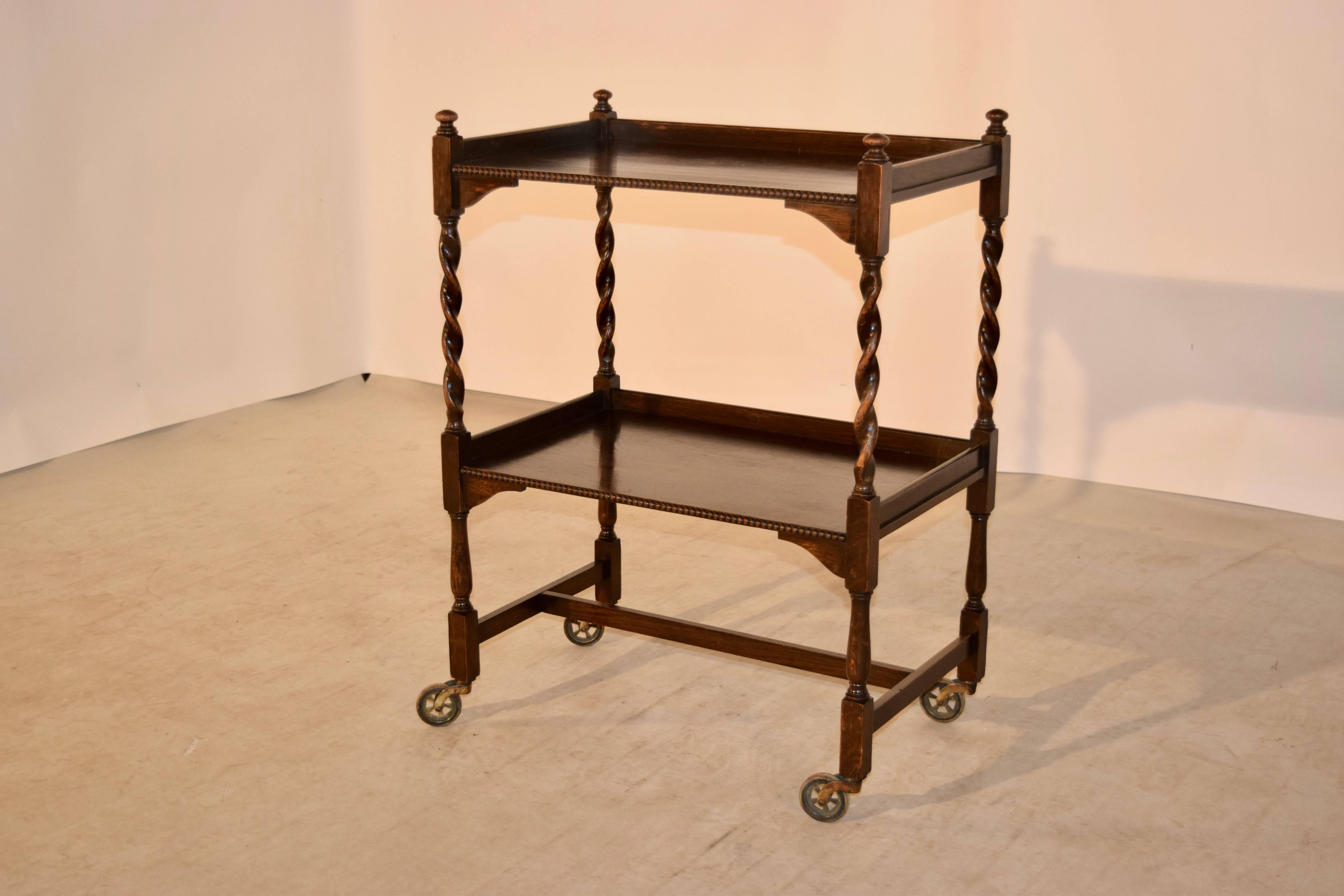 English drink cart made from oak, circa 1900. The top is decorated with finials, following down to two shelves, which have galleries around them and beaded fronts. The shelf supports are barley twist and have contrasting turned legs, joined by cross