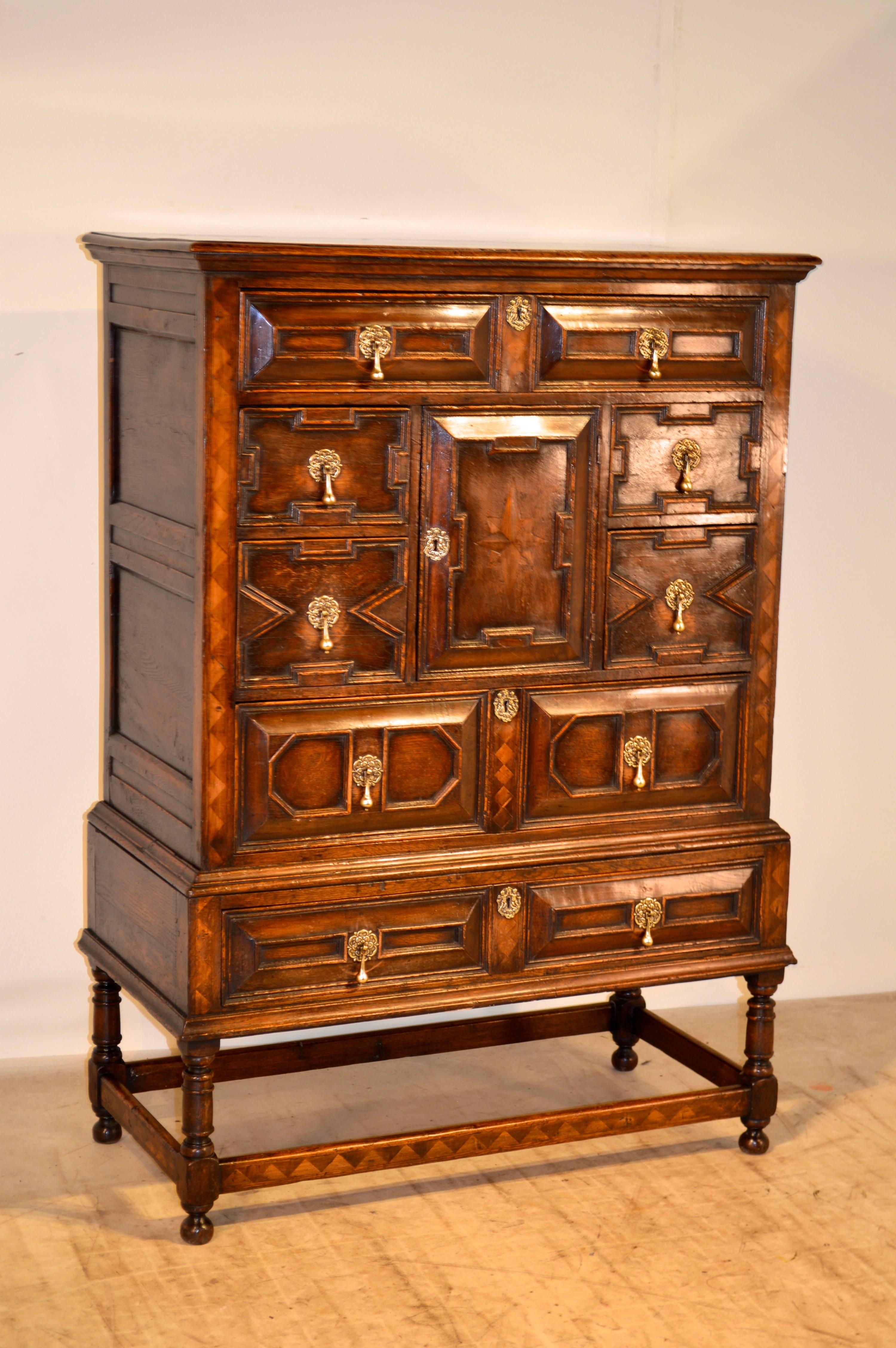 English oak chest on stand made from oak with walnut and satinwood accents, circa 1900. It has a single drawer over a central door flanked by two single drawers on each side, all over two long single drawers. The drawer fronts as well as the door