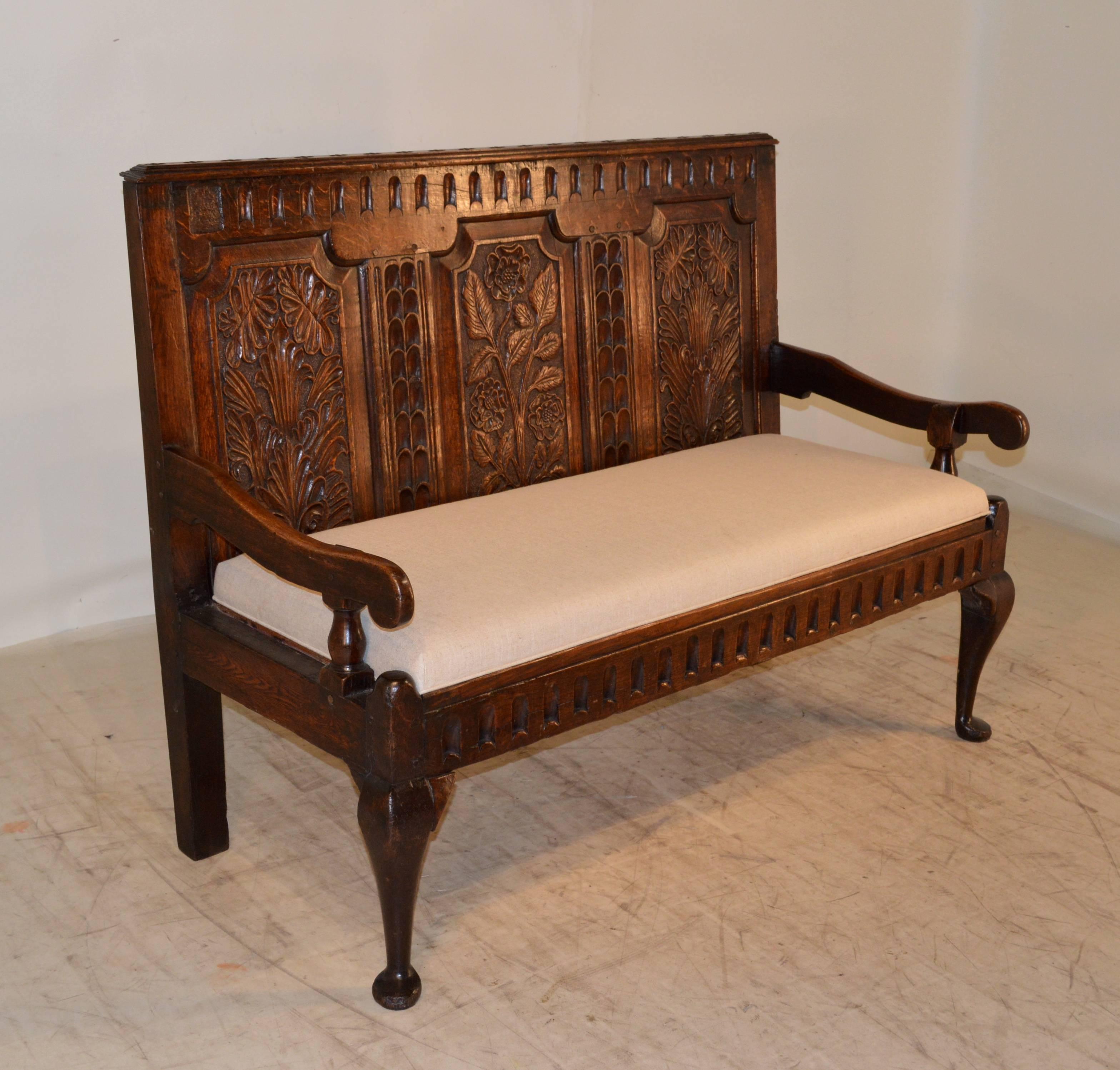 Early 18th century English oak carved settle with a raised and carved decorated paneled back.  The seat is made up of original oak slats, and we recently had it upholstered with cushioning in linen for comfort.  The apron is carved as well and is