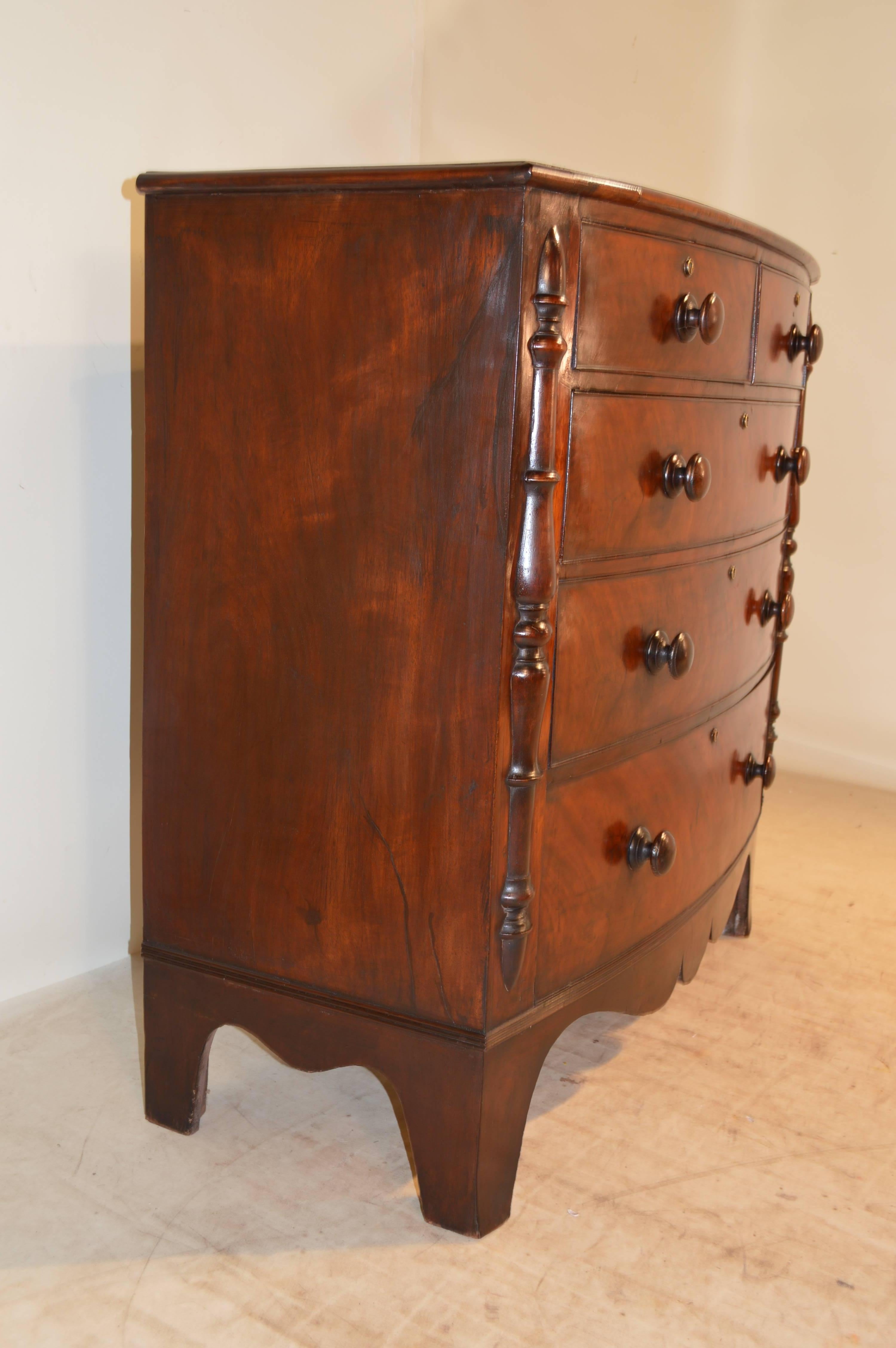 19th century English bowfront chest of drawers made from mahogany. The top has a beveled edge and follows down to a two drawer over three drawer configuration, all flanked by applied split column decoration. The skirt is scalloped and the case is
