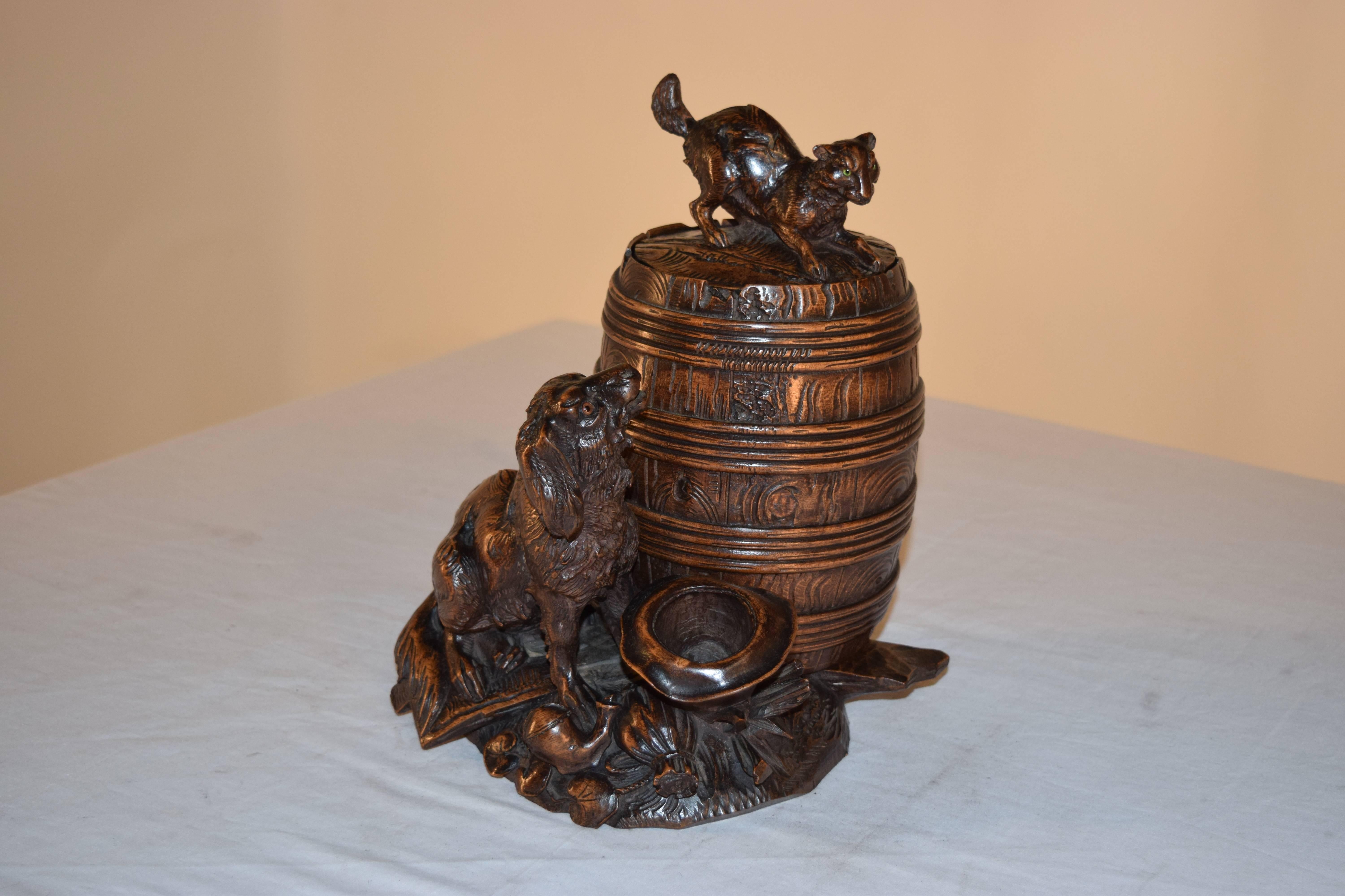 19th century hand-carved black forest humidor with a figure of a dog and cat. The cat is atop a barrel, which has a lid which lifts to reveal a compartment for tobacco. The barrel is sitting on a base with hand-carved sticks and logs and an upside
