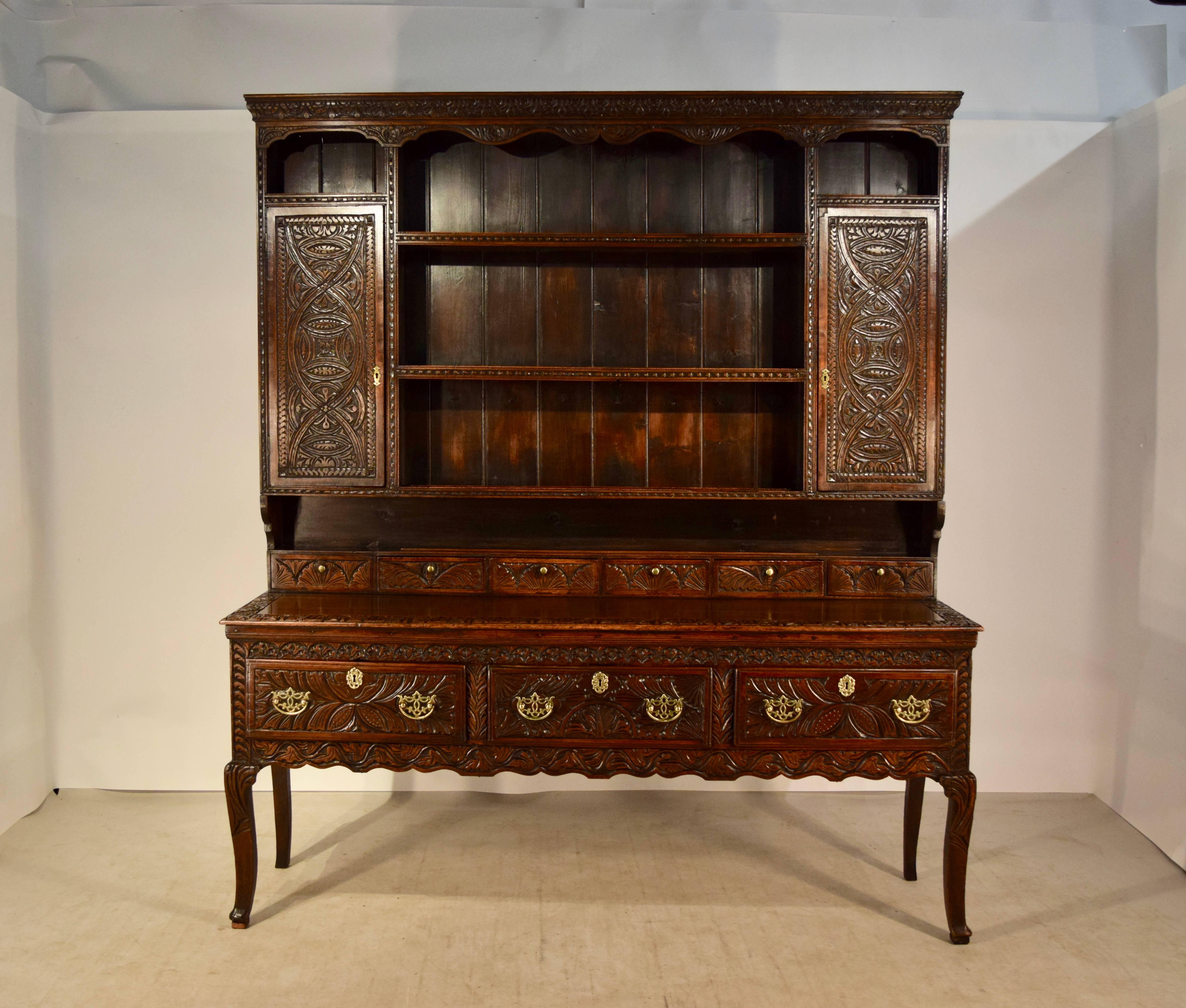 18th century oak dresser from Wales. The crown is carved decorated and has a scalloped apron over the top of the cupboard, which has small shelves over hand-carved doors, which are flanking a central set of three shelves, all with carved shelf