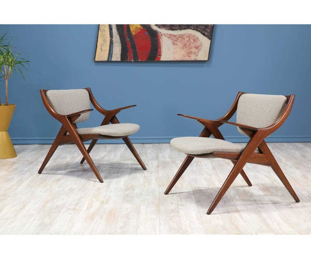 Mid-Century Modern scissor lounge chairs designed in the 1950’s. The sharp angled legs contrast nicely with the paddle arm rests. The frame is comprised of walnut-stained mahogany wood while the seatrest and backrest are upholstered in a