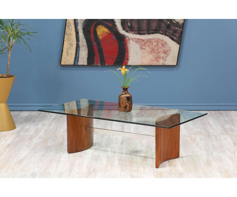 Mid-Century Modern coffee table designed by German-born architect and furniture designer Vladimir Kagan for Selig in the 1960’s. With a wide chrome steel bar stretching across a two-part walnut base and a new, rectangular glass top, its curved and