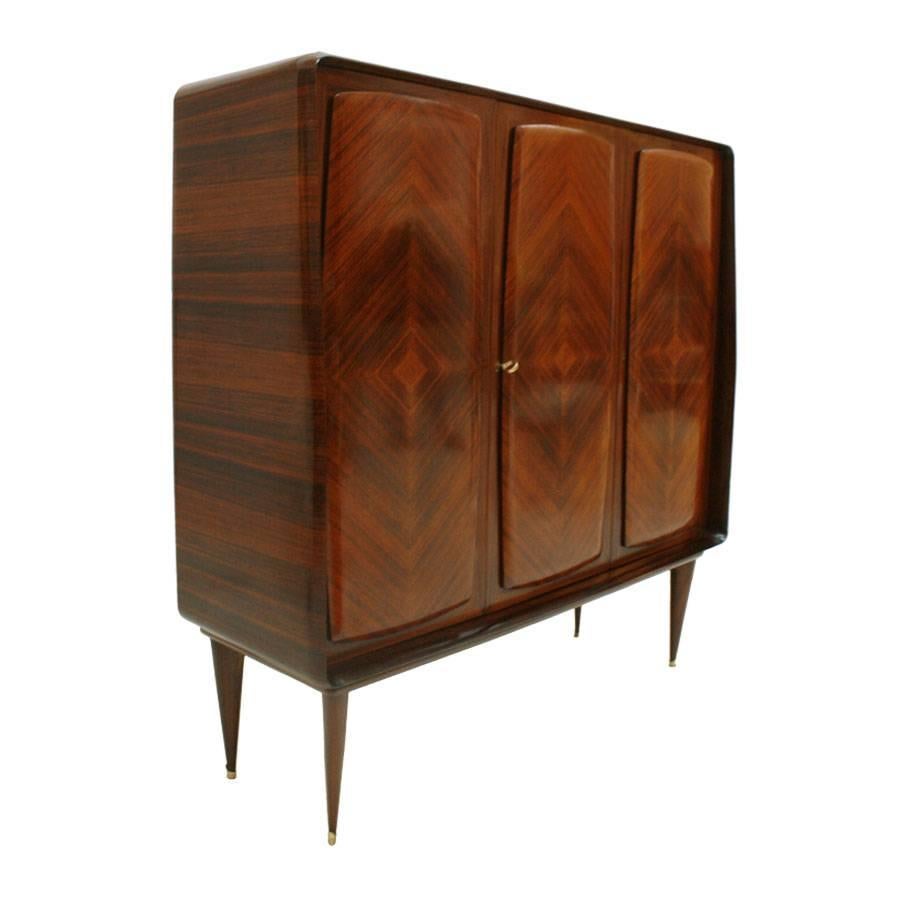 Three-door bar cabinet with drawers inside, made in solid wood clad in rosewood. Doors and marquetry made of rosewood. Legs with brass finish.