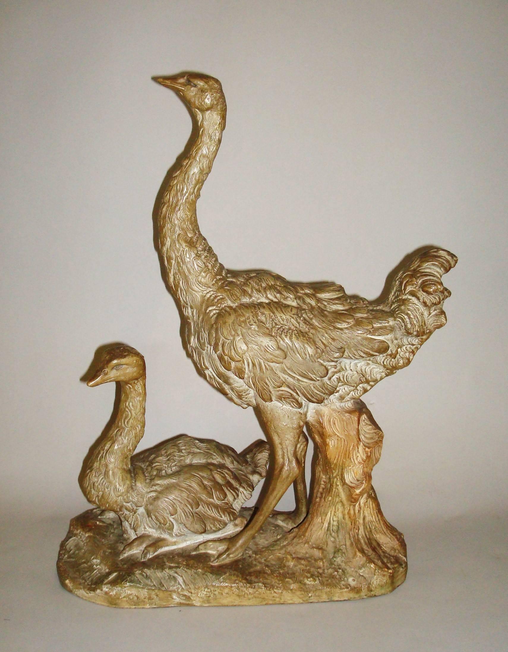 Early 20th century terracotta sculpture of two ostriches with a patinated bronzed finish. The larger ostrich, poised with head raised and prominent tail feathers, the smaller bird, presumably her chick sitting at her feet. The whole on an oval