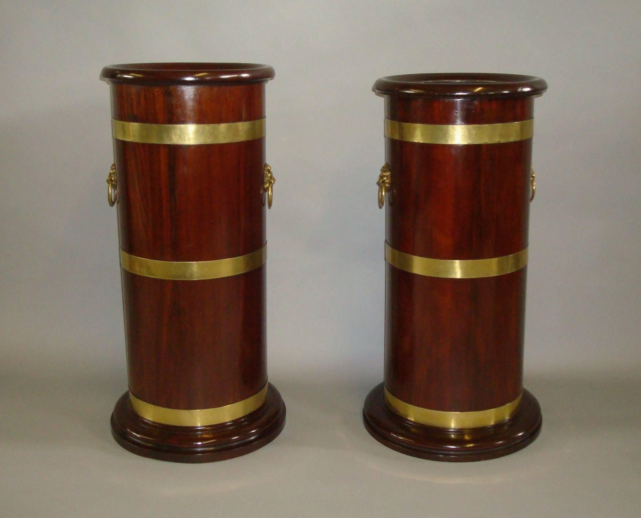 Late 19th century matched pair of mahogany and brass stick or umbrella stands of cylindrical form. The moulded shaped rims overhanging the cylindrical bodies which have three brass bound straps and brass lion mask handles to either side, raised on