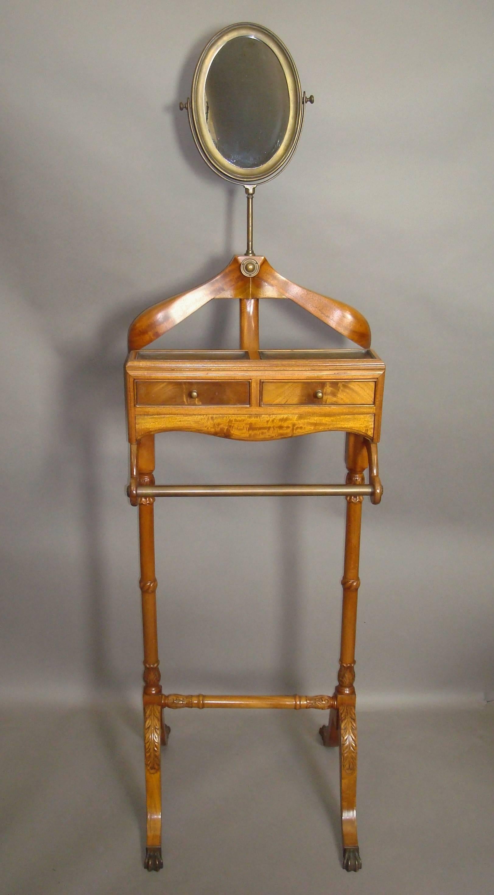 Good 20th century figured mahogany gentleman's valet stand; the swivel oval mirror in a brass moulded frame, raised on a brass support above the substantial shaped jacket hanger. The main compartment with two small drawers and two brass dished trays