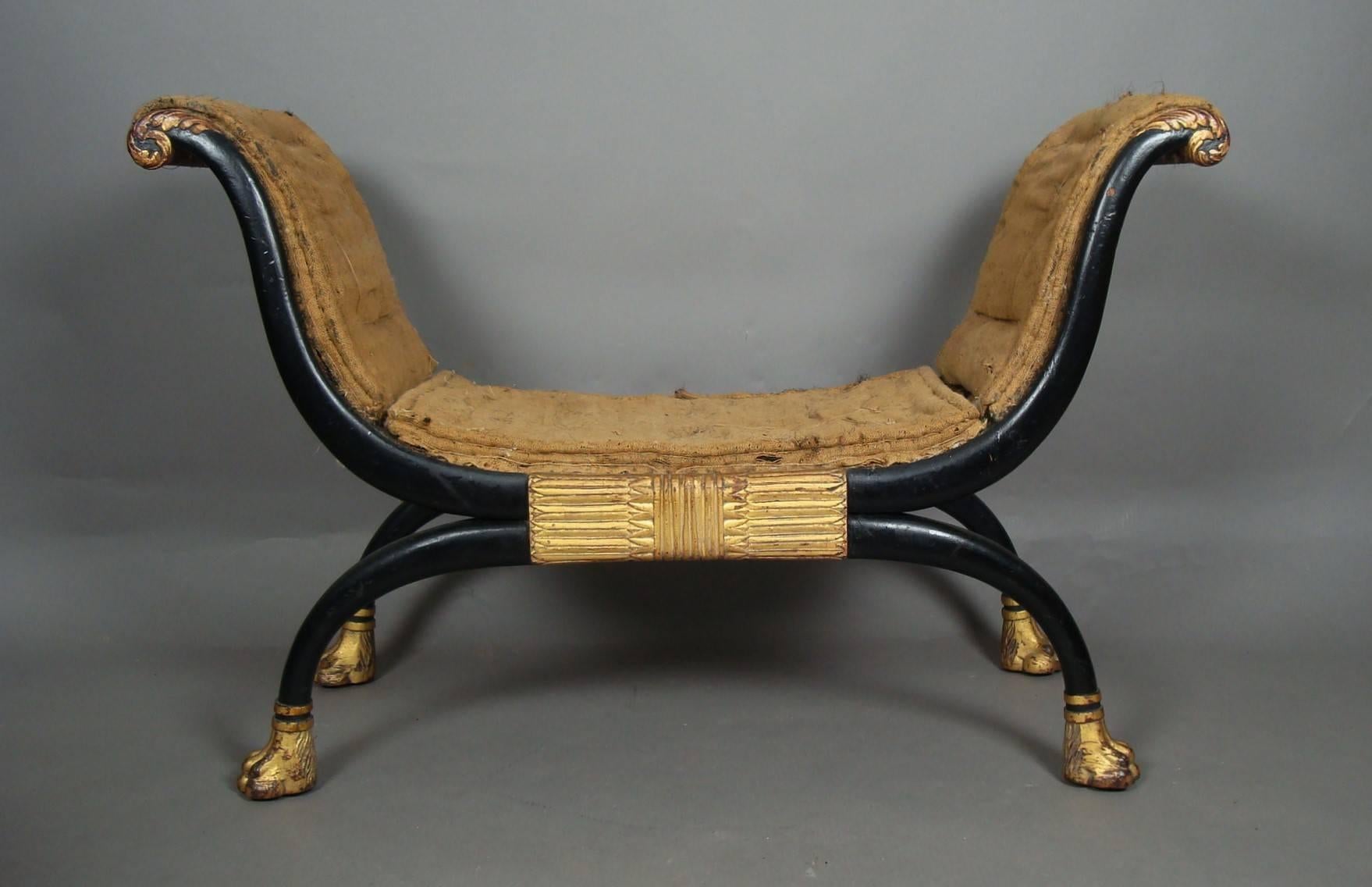 An exceptional Regency window seat, in the manner of Thomas Hope; the ebonised exaggerated cross frame, headed with carved giltwood acanthus scroll decoration. The central large carved tablet of classical design. The scroll legs with giltwood